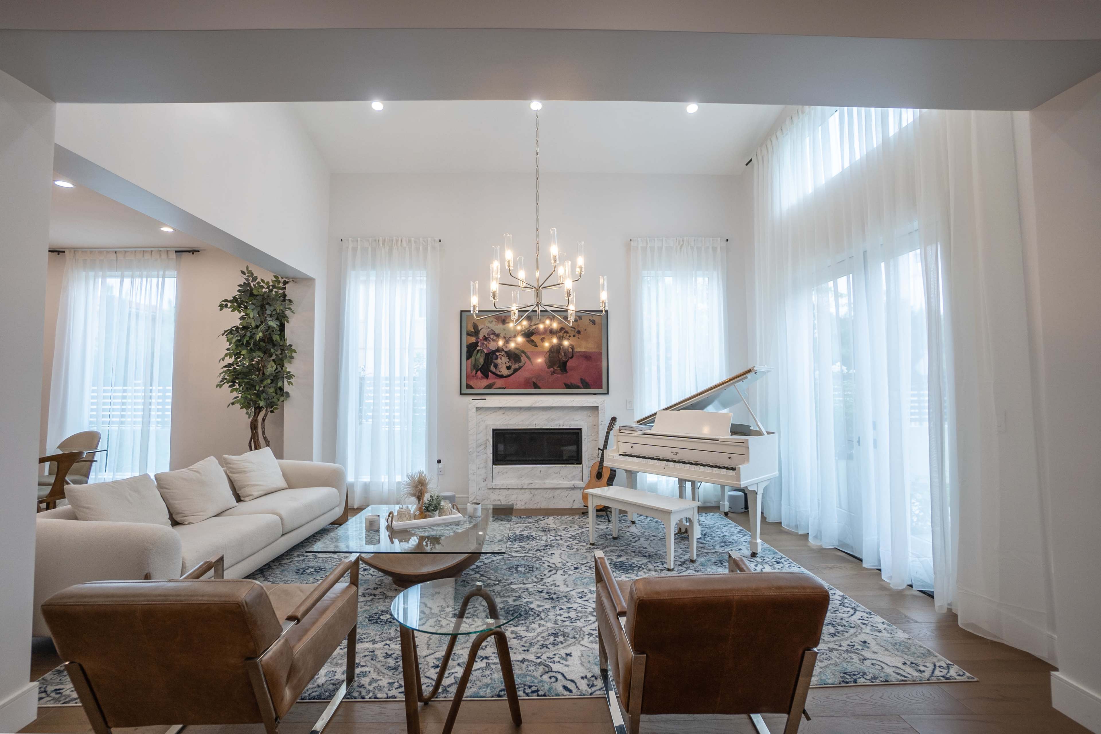 A bright living room features a white sofa, two brown armchairs, a glass coffee table, and a grand piano, with large windows draped in sheer curtains.