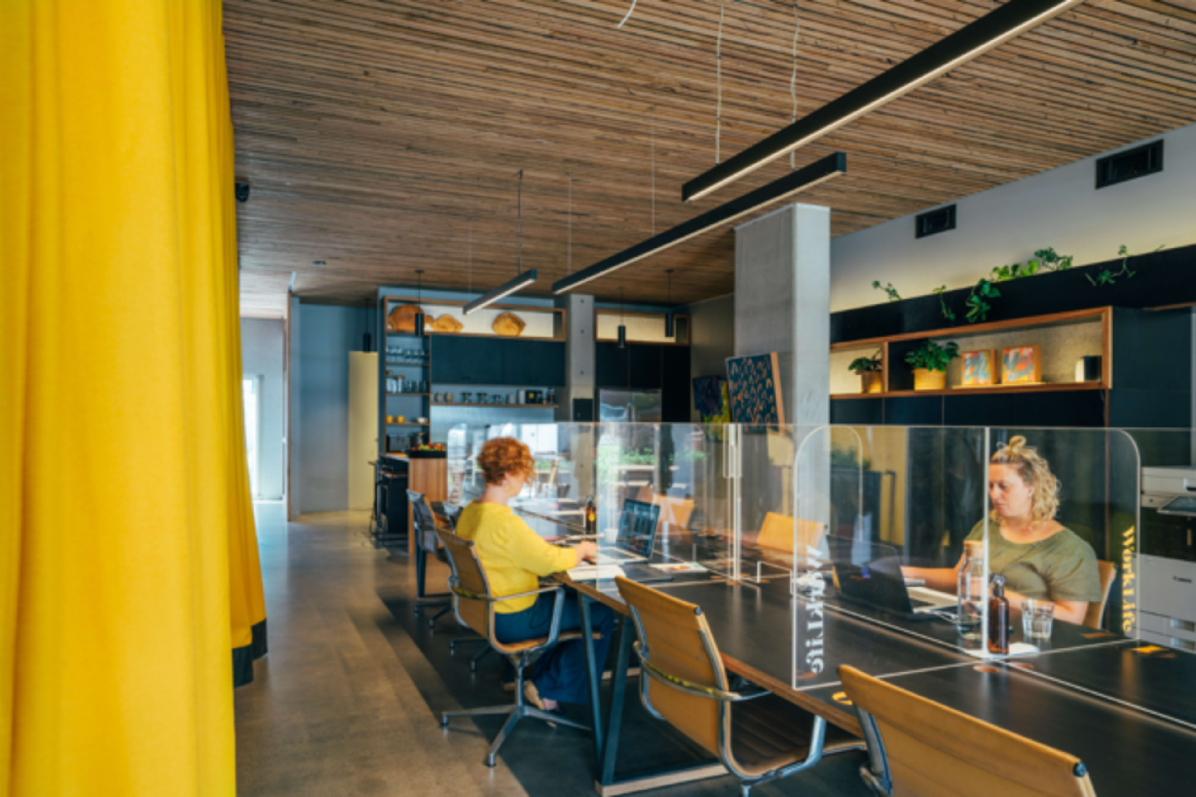 A modern workspace with wooden ceiling beams and glass partitions, featuring two women working at desks.