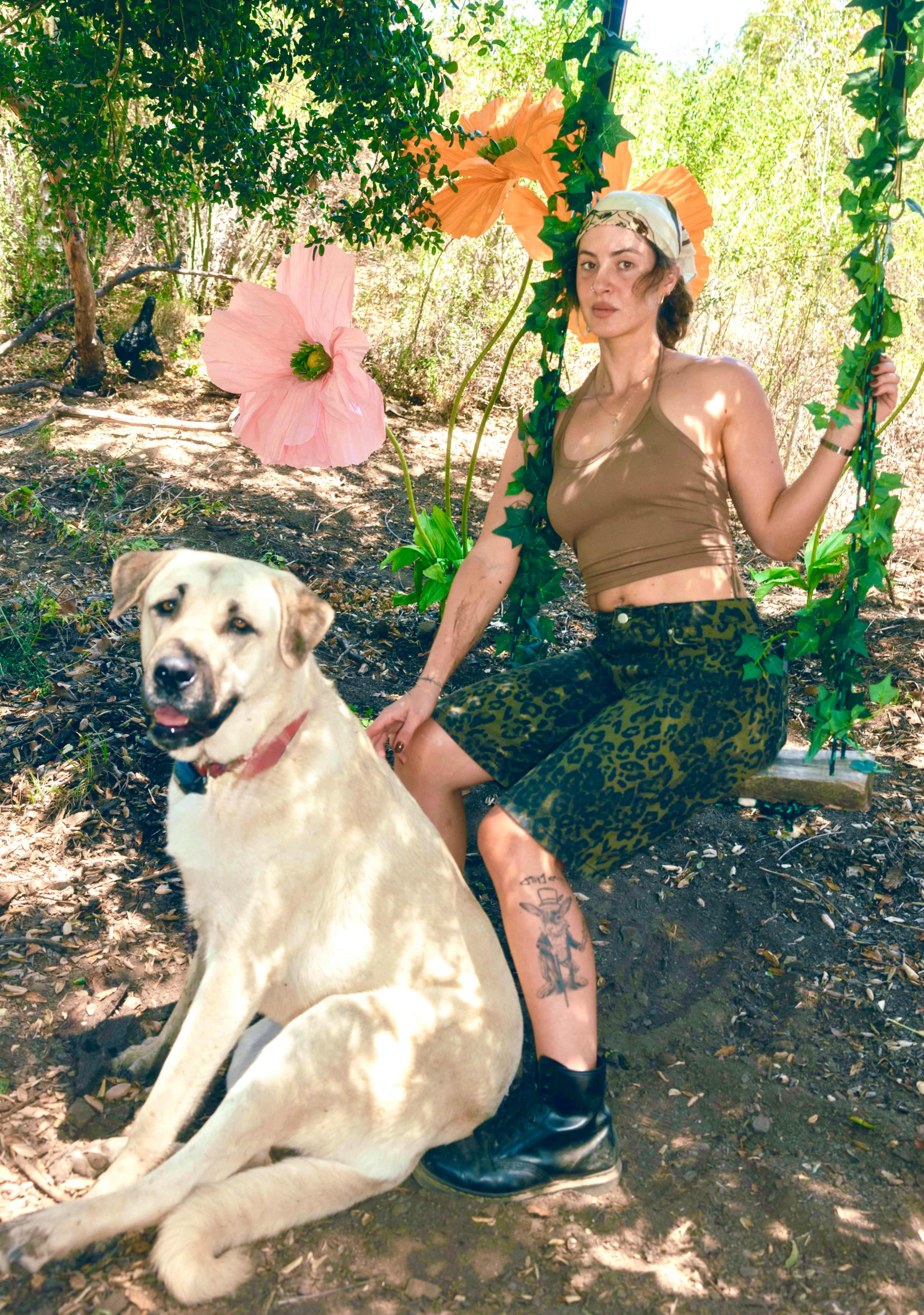 A woman sits on a swing beside a large light-colored dog, surrounded by greenery and oversized flowers.
