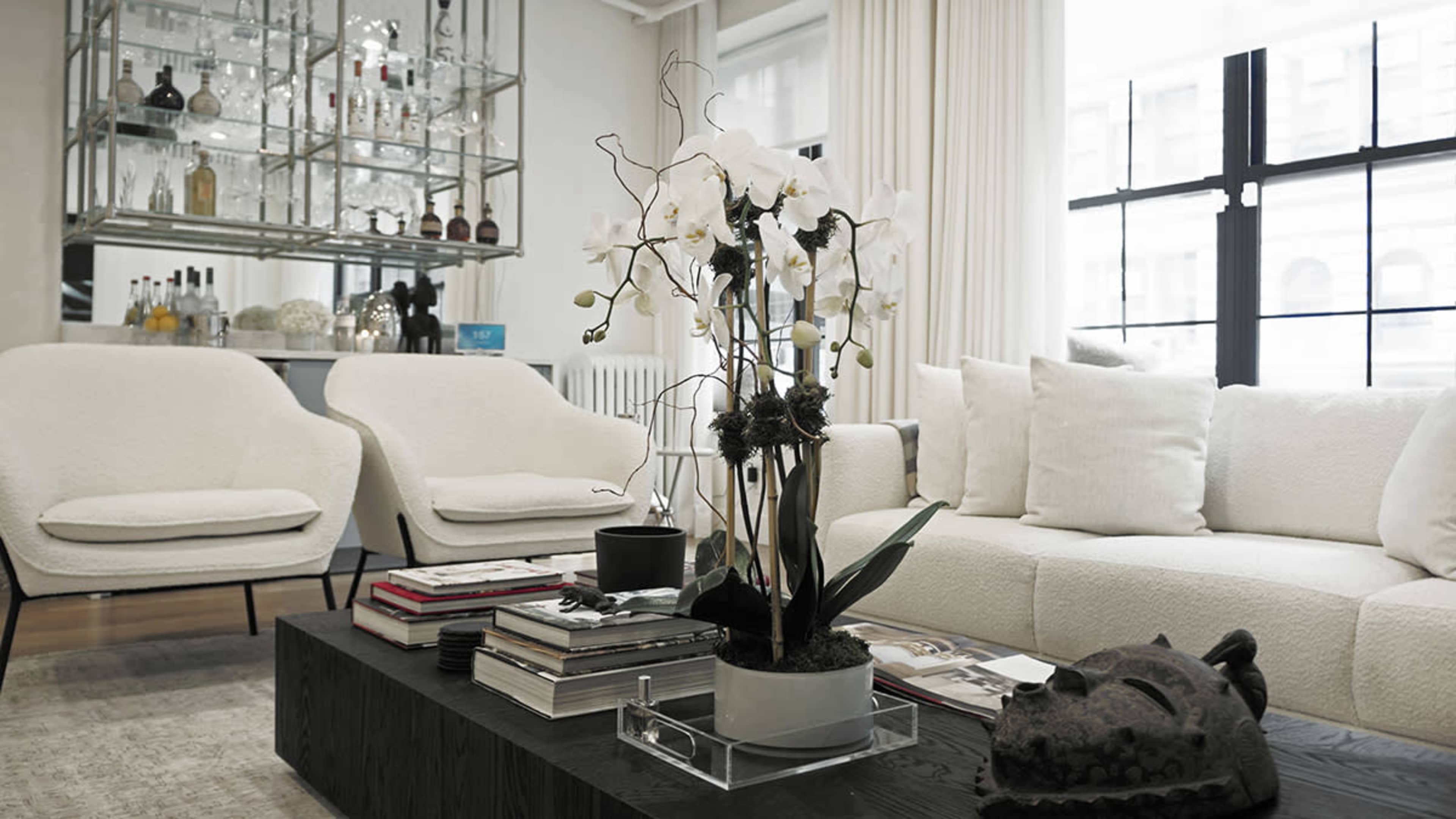 The image shows a modern living room featuring two white armchairs, a large white sofa, a coffee table stacked with books, and a decorative plant with white orchids.