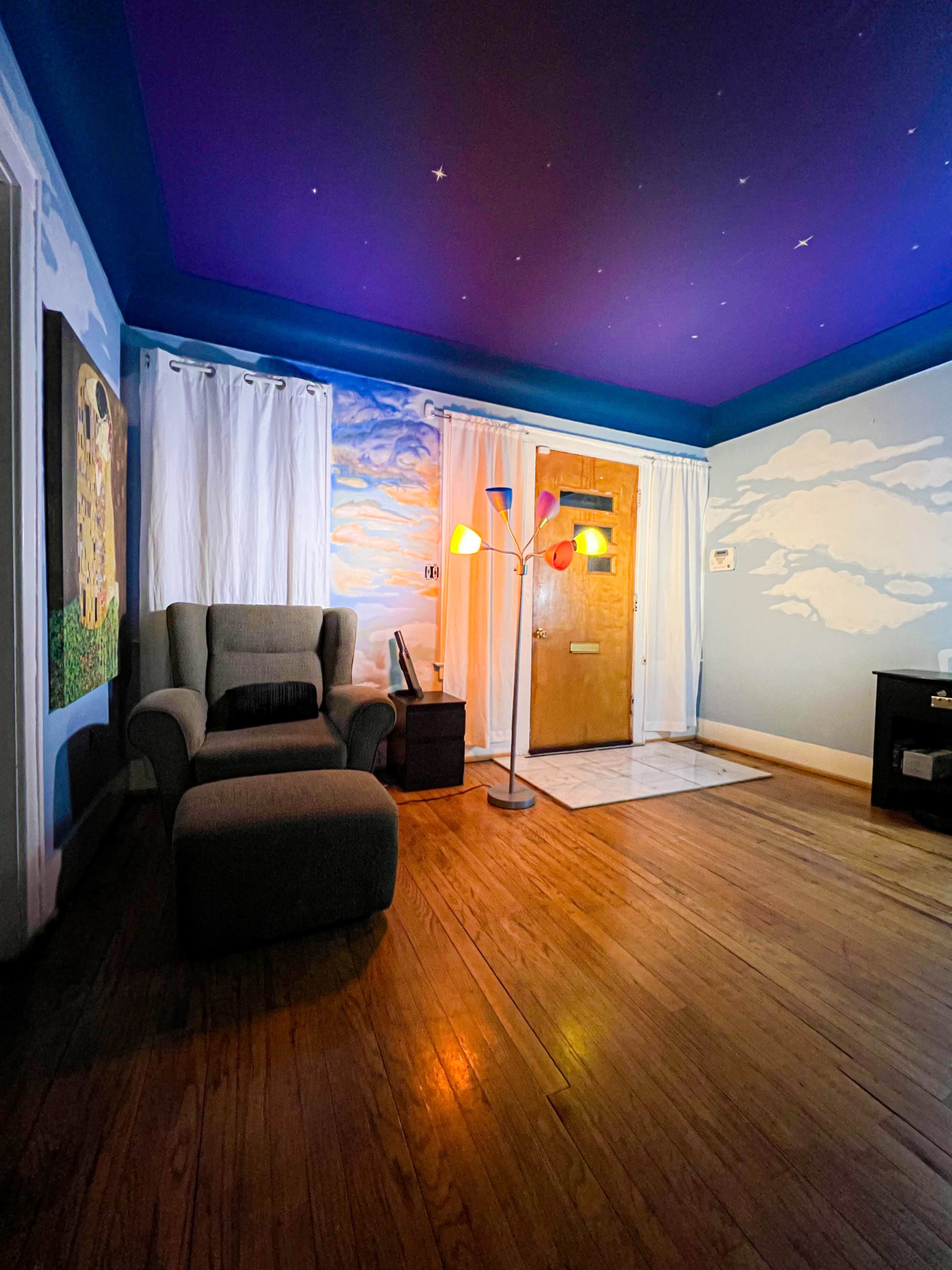 A living room features a wooden door, colorful murals of clouds on the walls, and a chair beside a floor lamp in the corner.