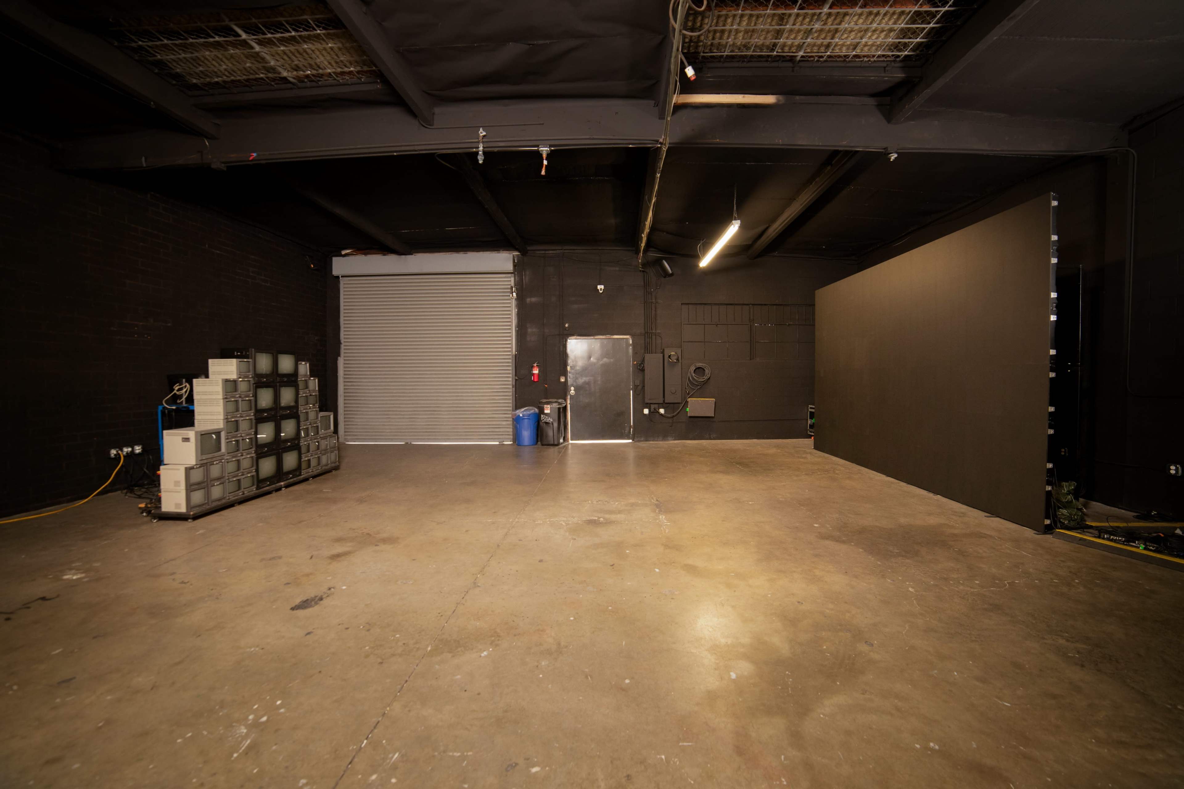 A large, empty industrial space with a concrete floor, a metal roll-up door, and several gray storage units against a black wall.
