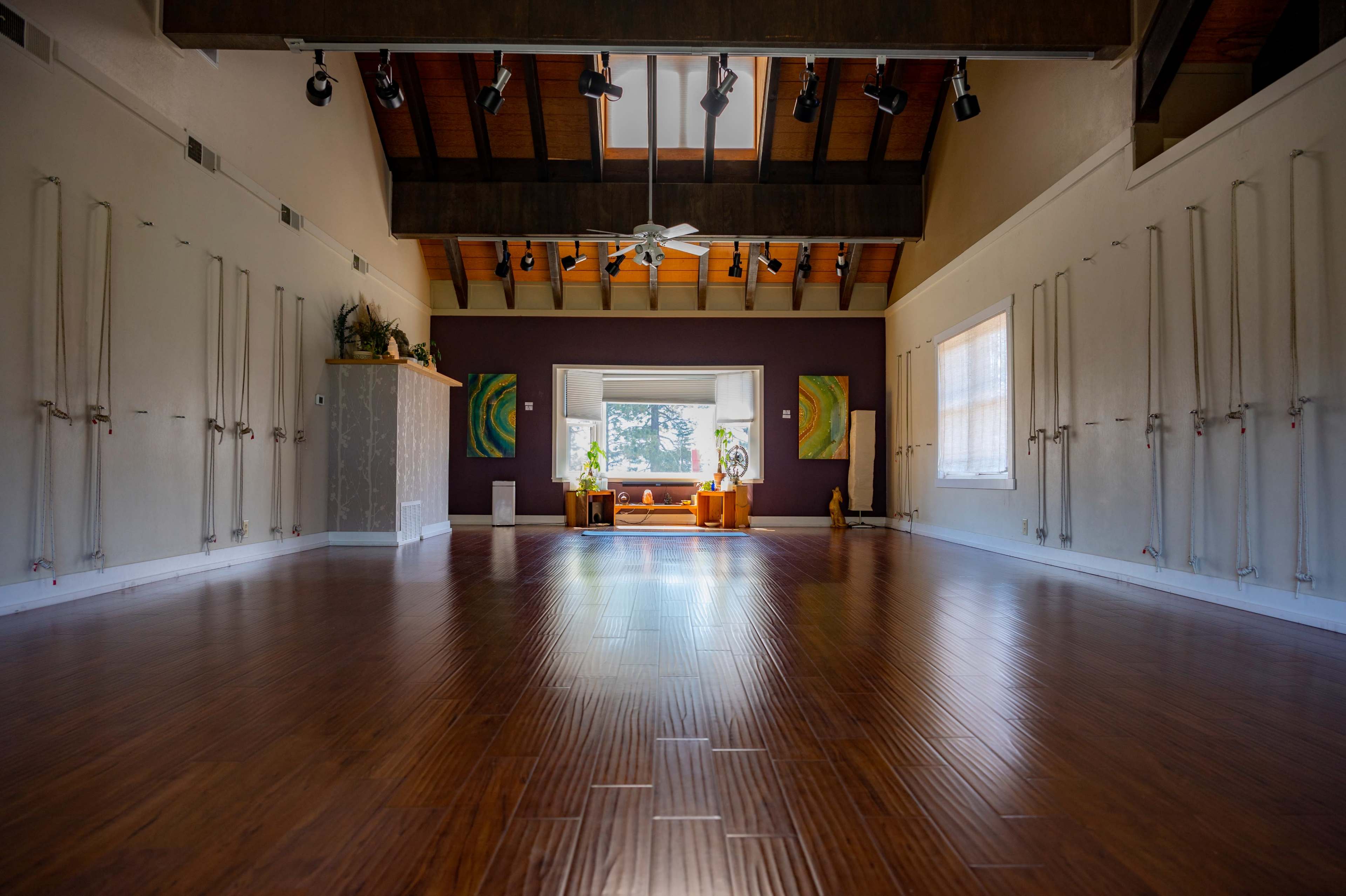 The image shows a spacious yoga studio with wooden floors, high ceilings, and large windows illuminated by natural light.