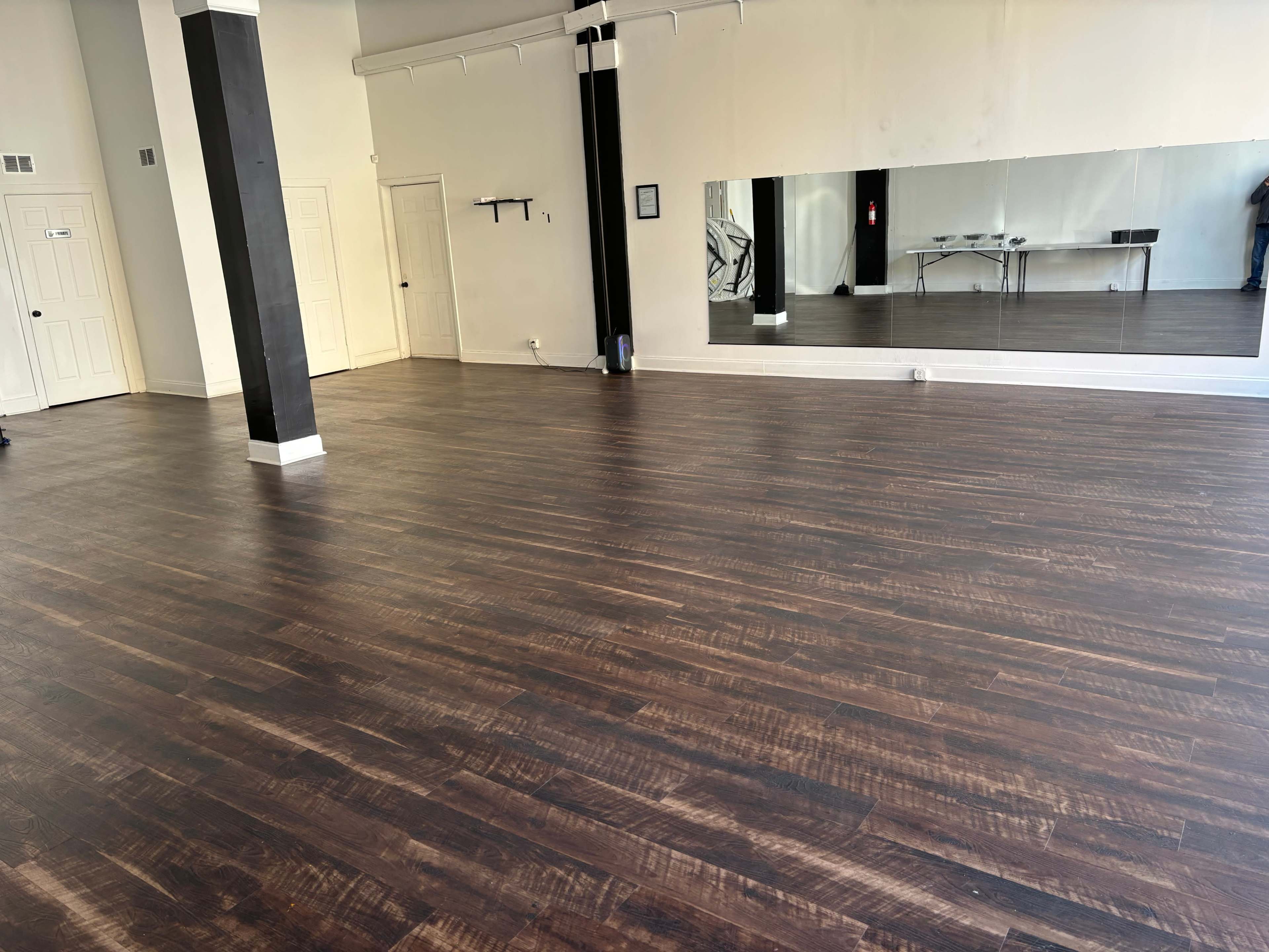 A spacious, empty room with dark wooden flooring, white walls, and a large mirror on one side.