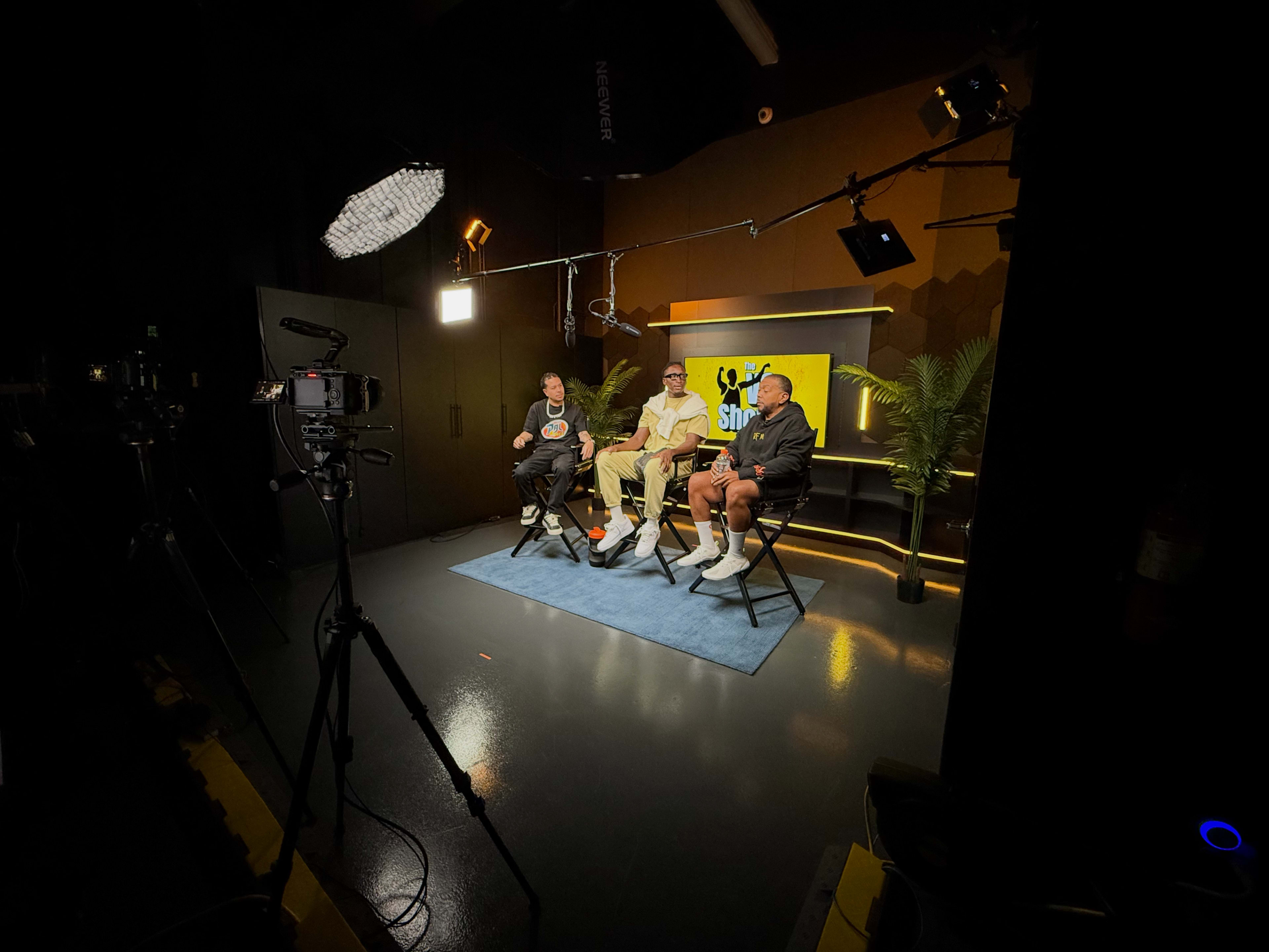 Three individuals sit on stools in a studio setting, facing the camera, with lighting equipment and a backdrop featuring a logo.