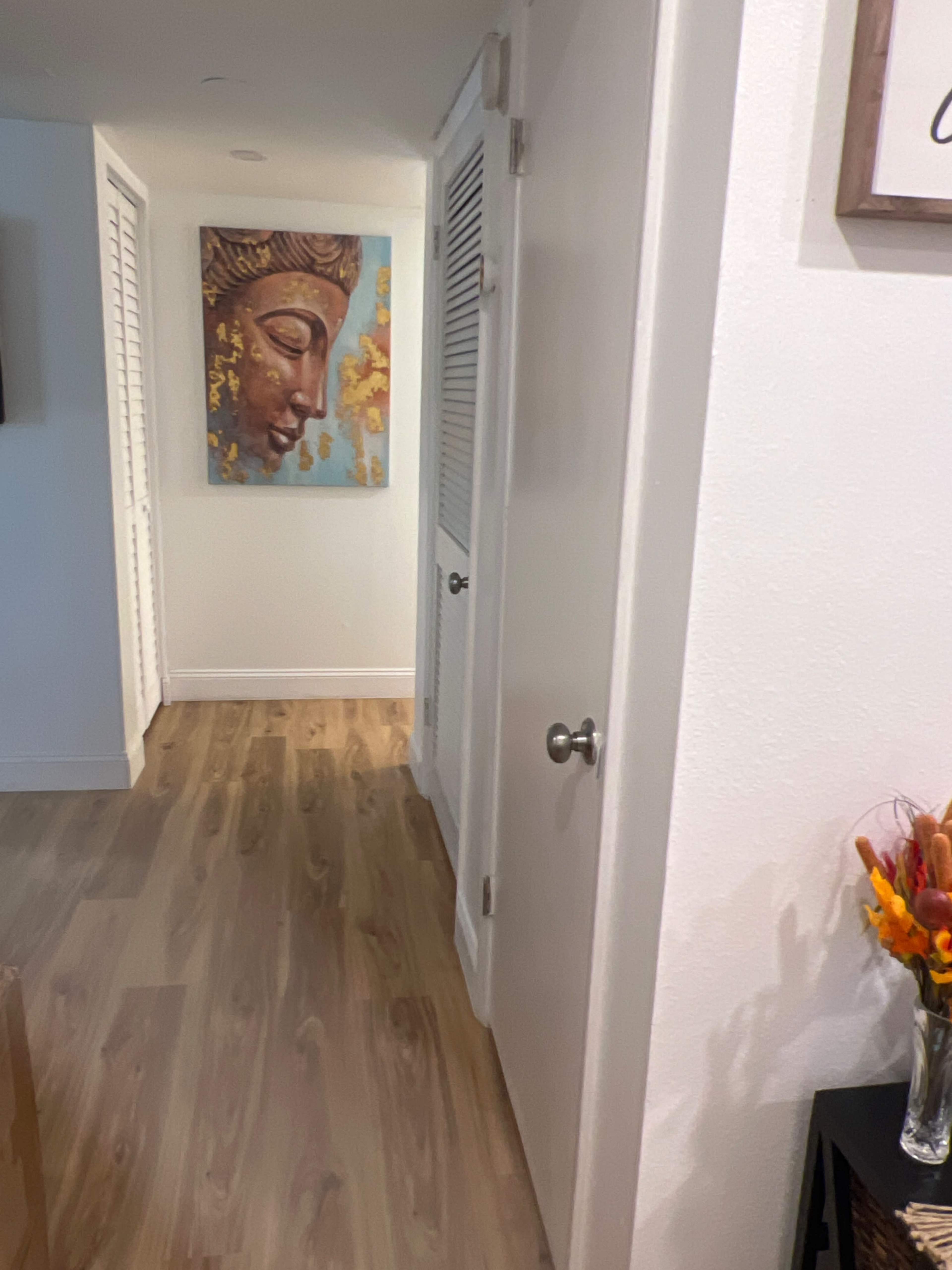 A hallway features a decorative Buddha painting on the wall opposite two closed doors and a wooden floor.