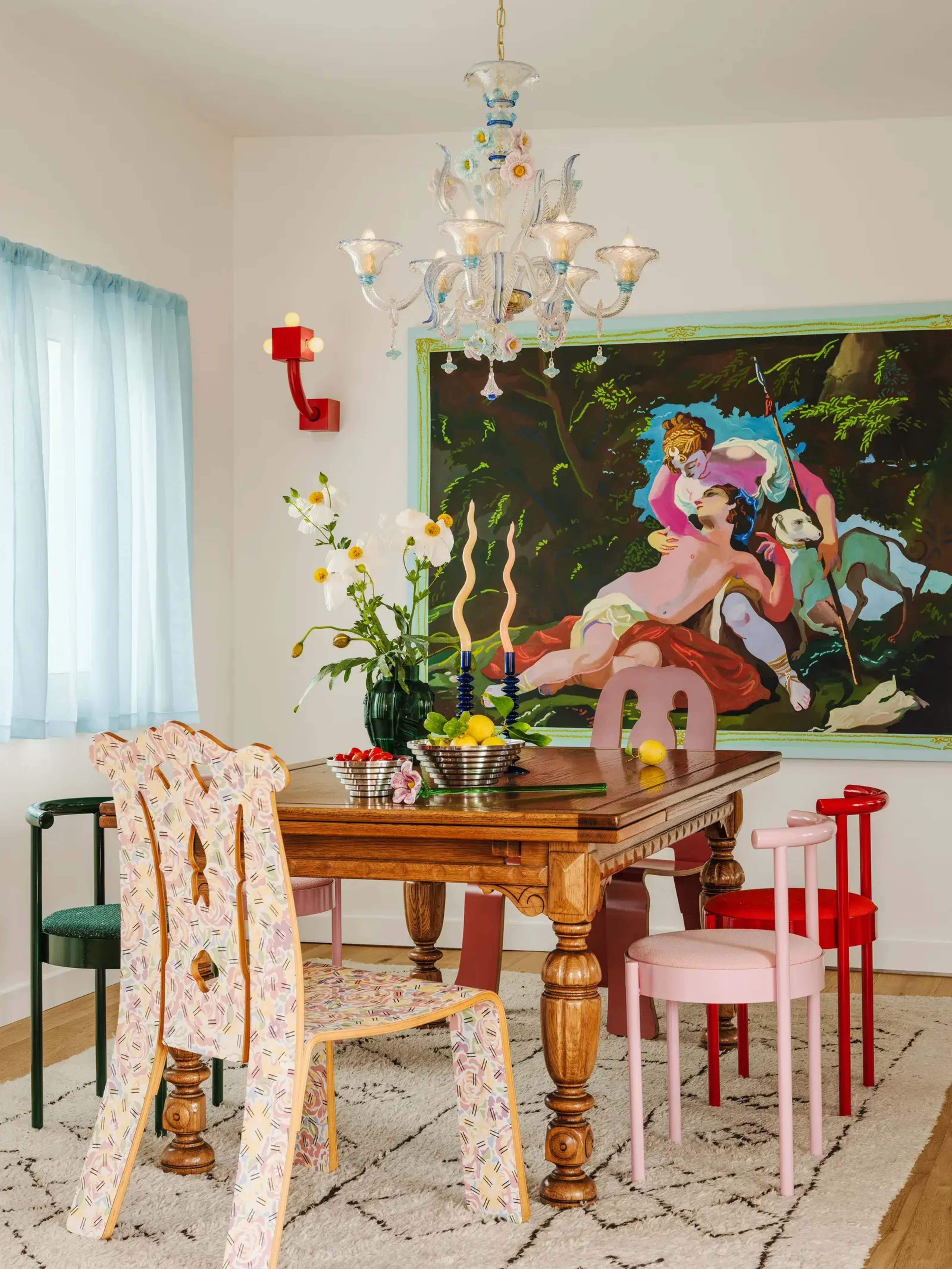 A dining area features a wooden table surrounded by colorful chairs, with a chandelier overhead and an artwork depicting figures on the wall.