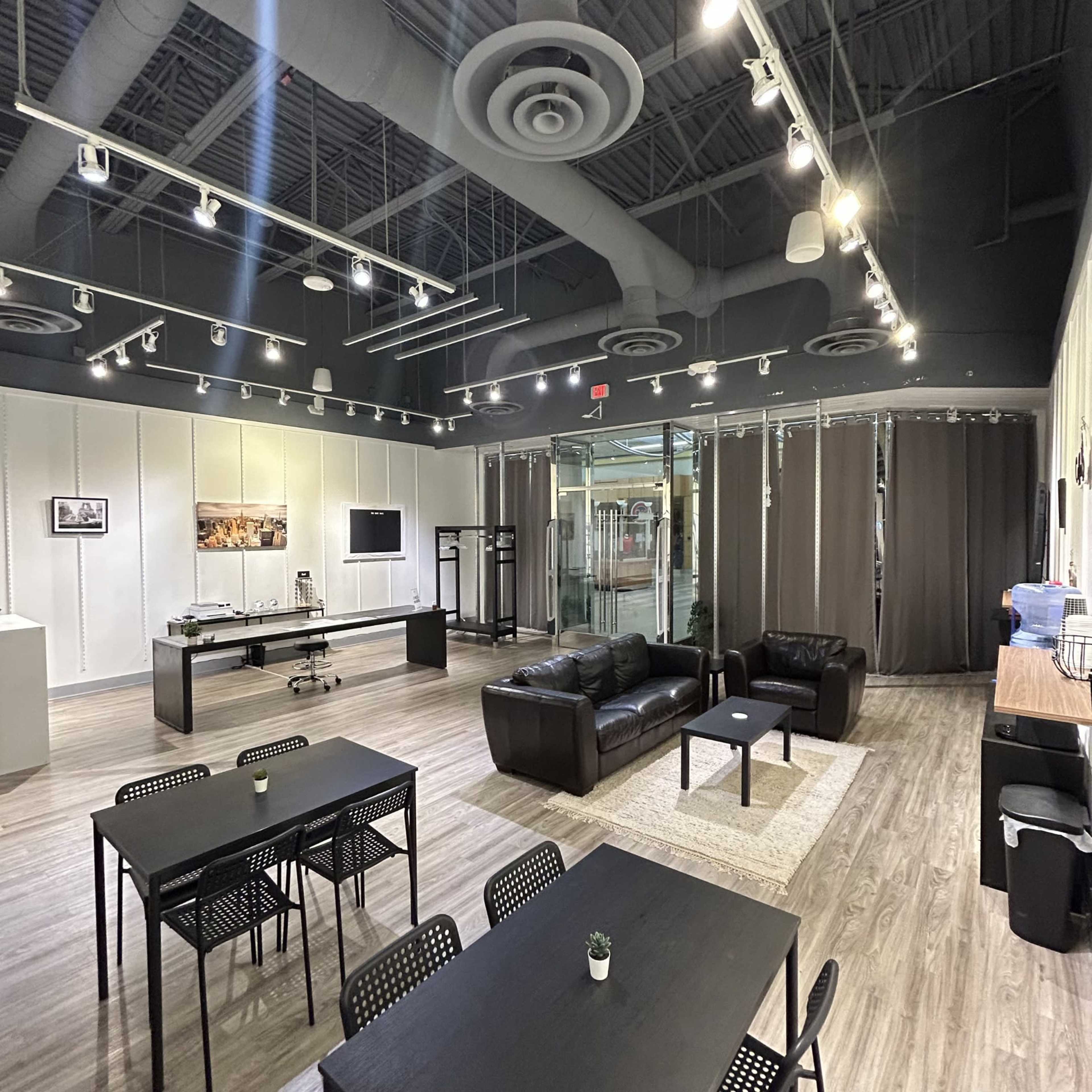 The image shows a modern, spacious room with dark furniture, including two couches and several tables, along with large windows and track lighting.