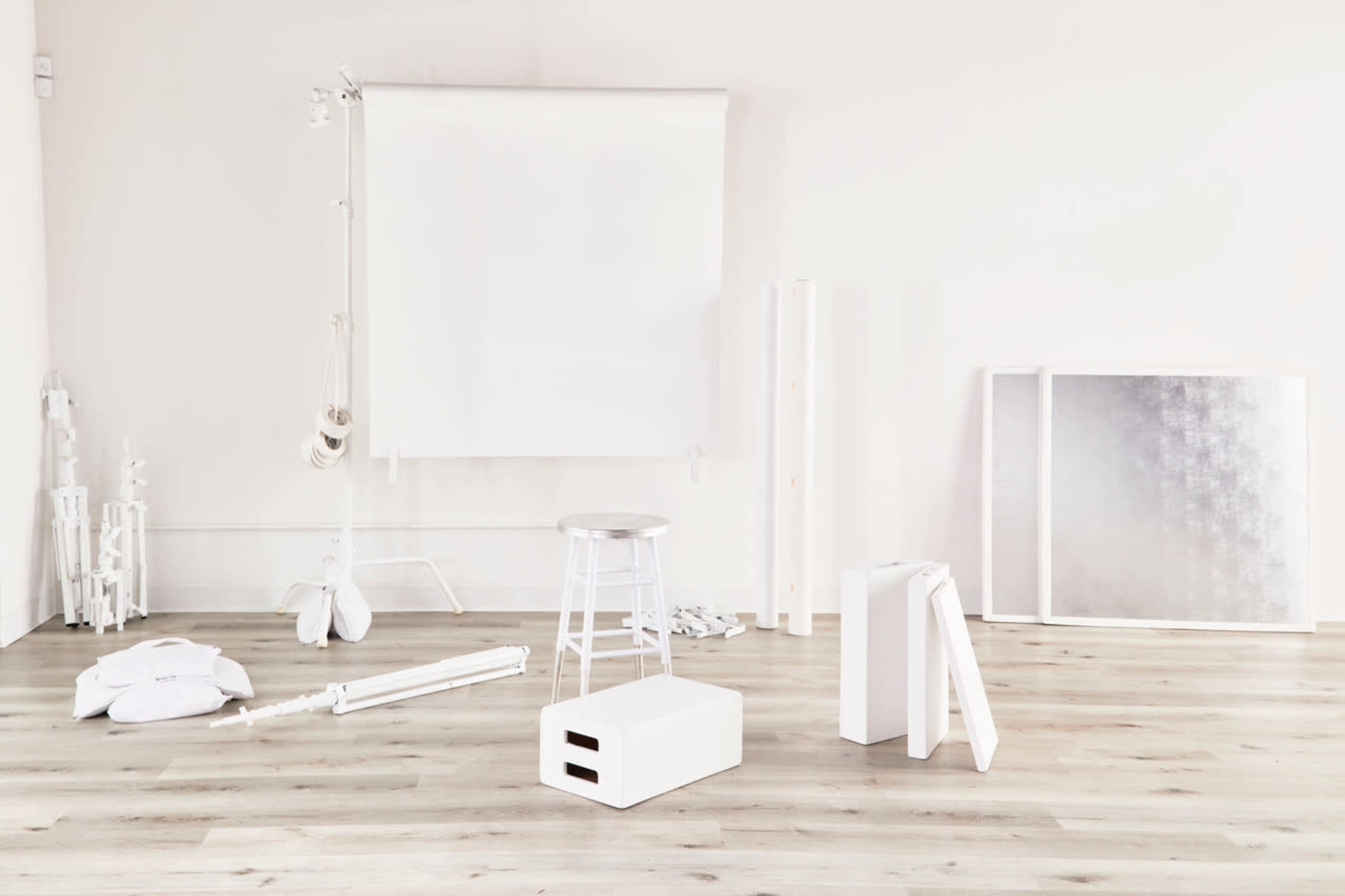 A minimally furnished studio space with various white objects, including a stool, a box, and backdrop equipment, set against a light wooden floor.