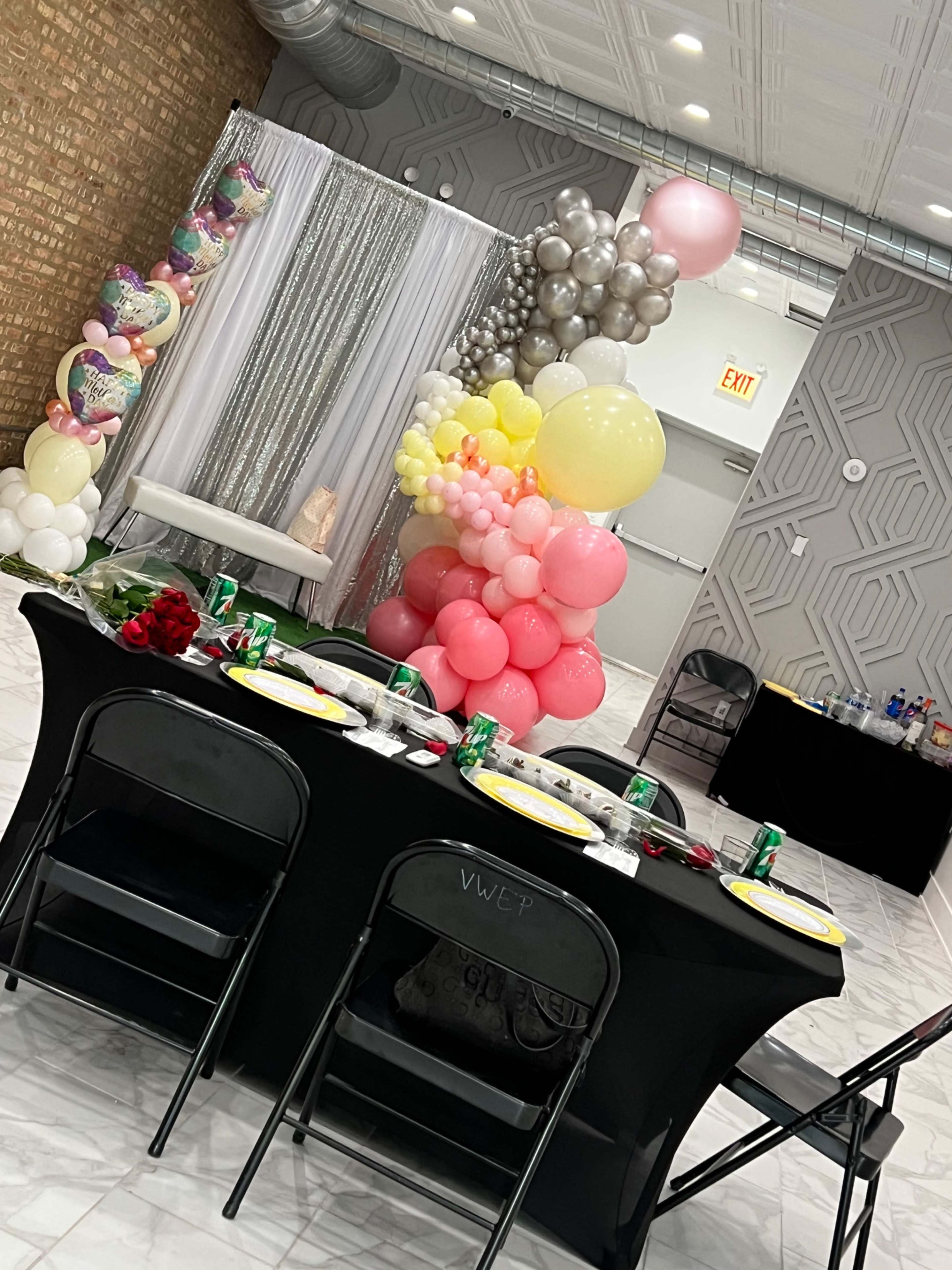A decorated event space features a table set with plates and drinks, surrounded by a backdrop of balloons and a shimmering curtain.