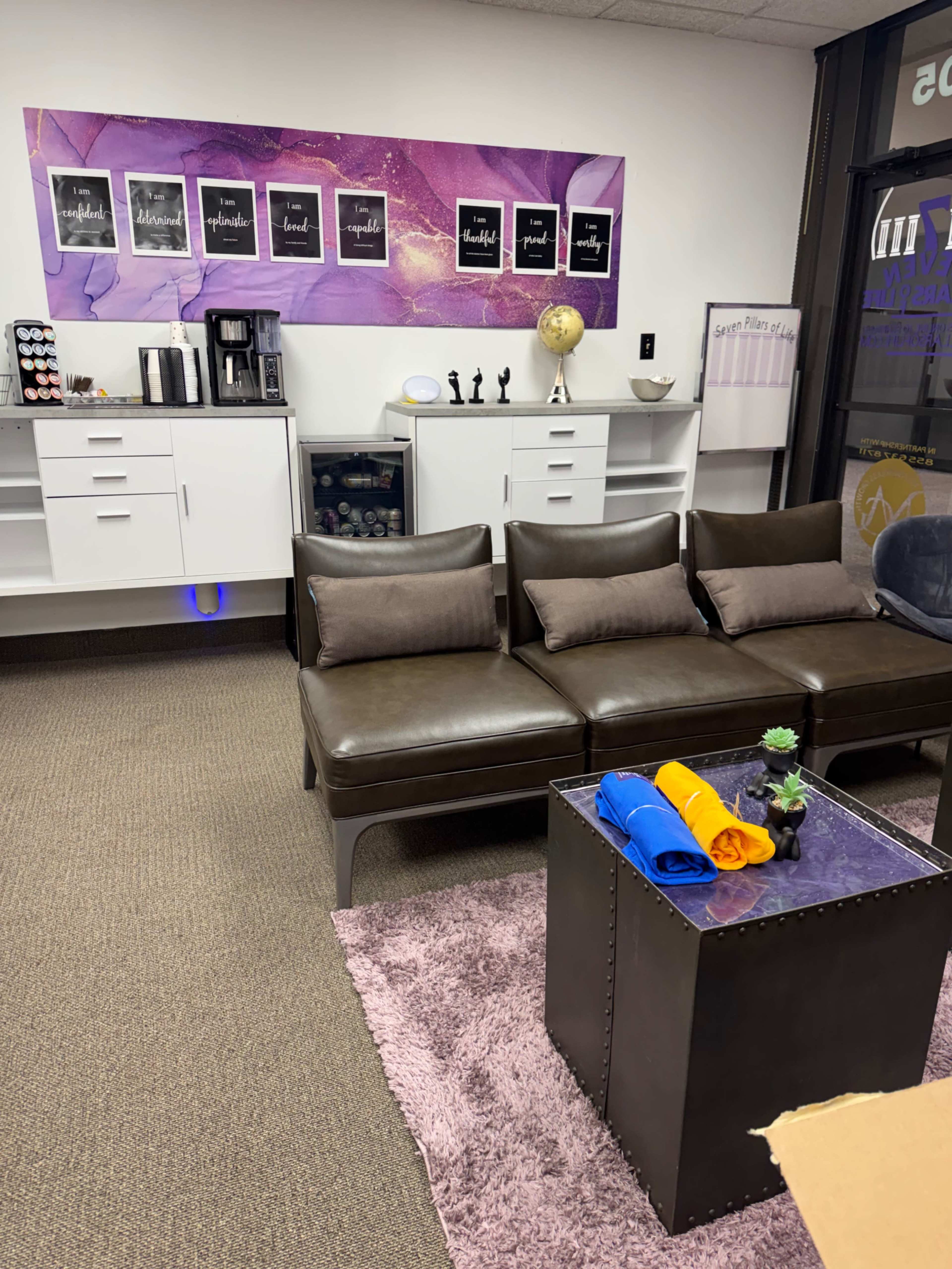 The image shows a modern office space with a brown leather sectional couch, a black metallic coffee table, and a wall decorated with framed words, alongside a coffee station and a globe.