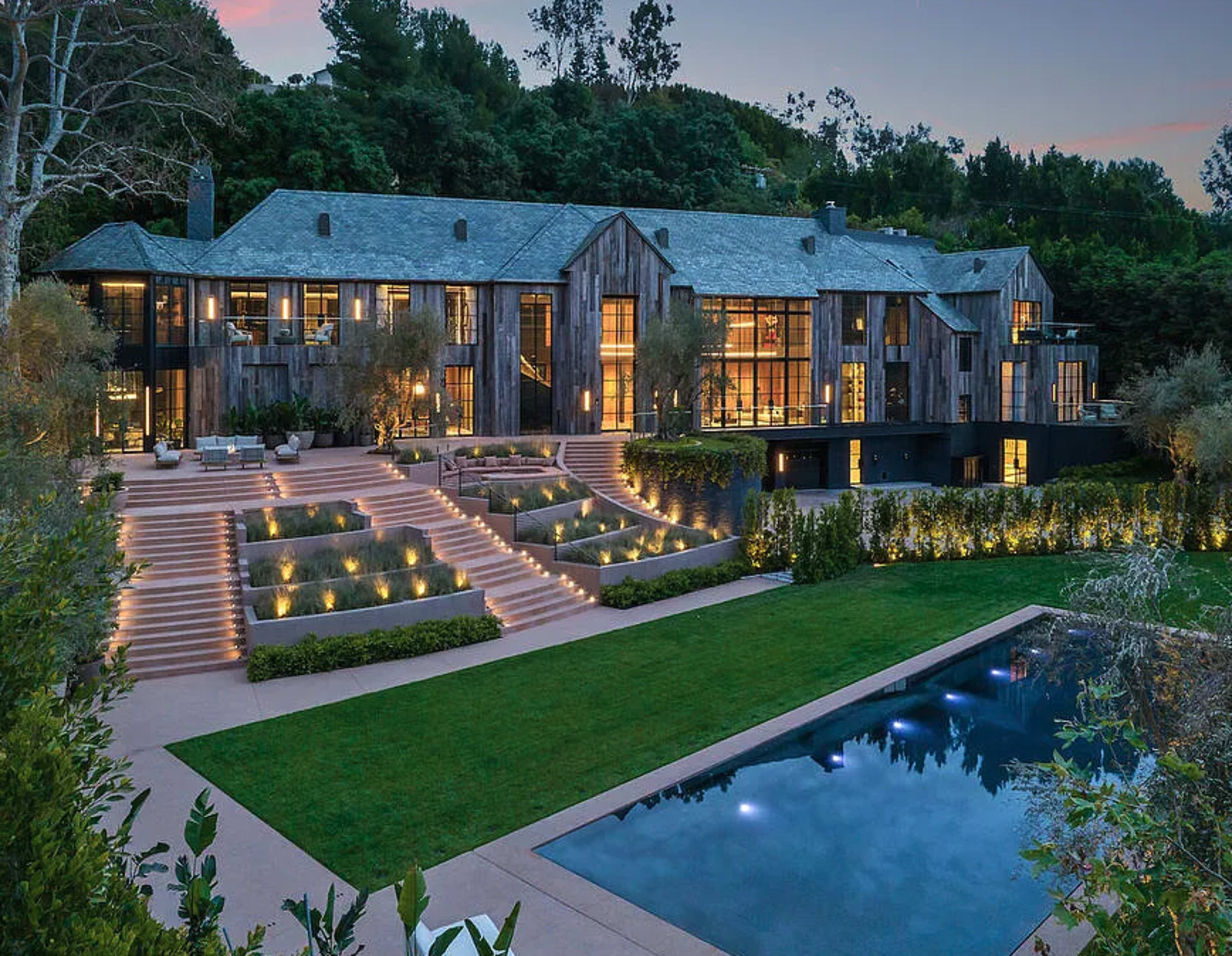 A large, modern house with multiple levels and glass facades is surrounded by landscaped gardens and an outdoor pool.