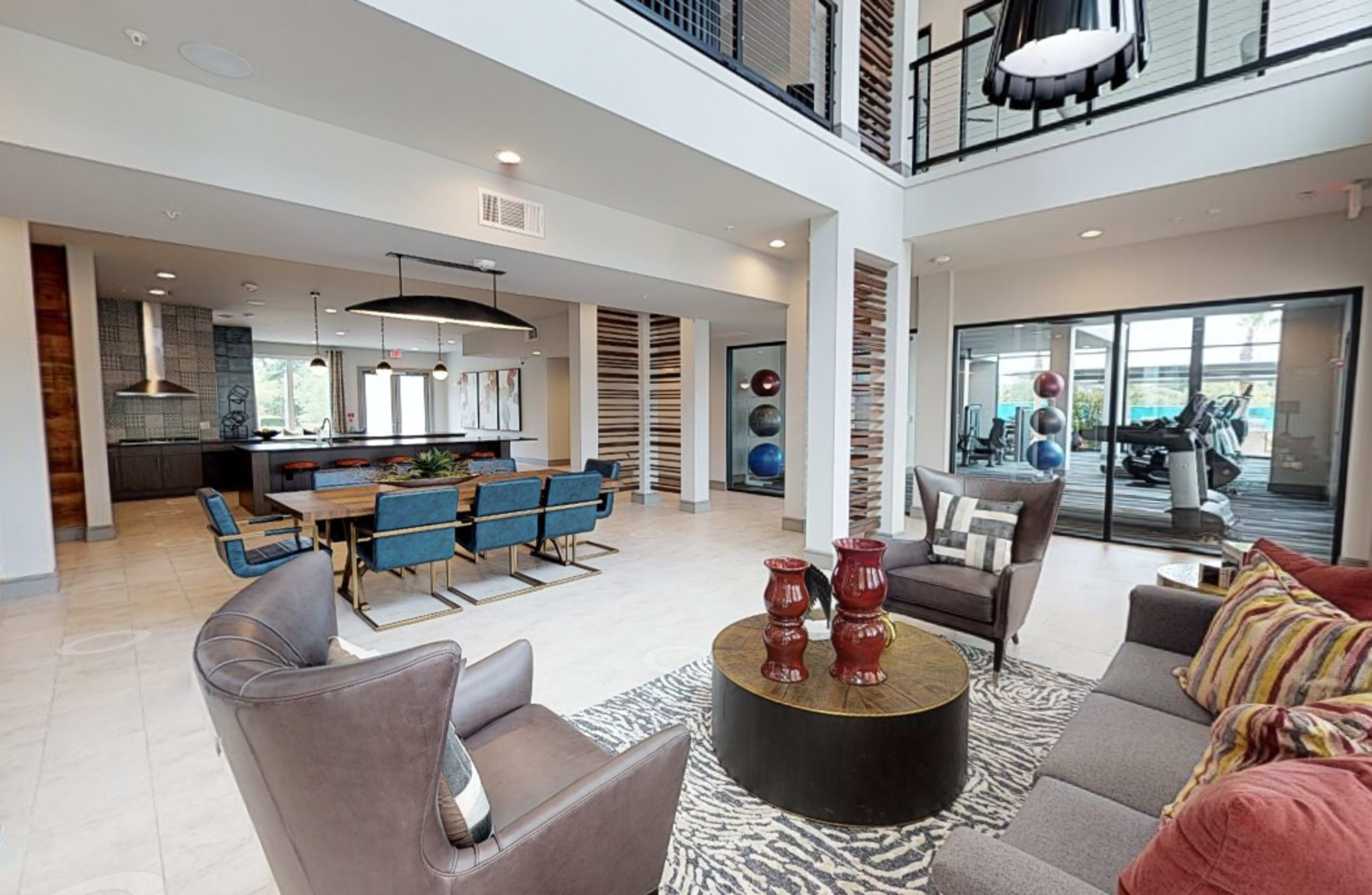 A modern communal lounge area with seating, a dining table, and an adjacent fitness room visible through large windows.