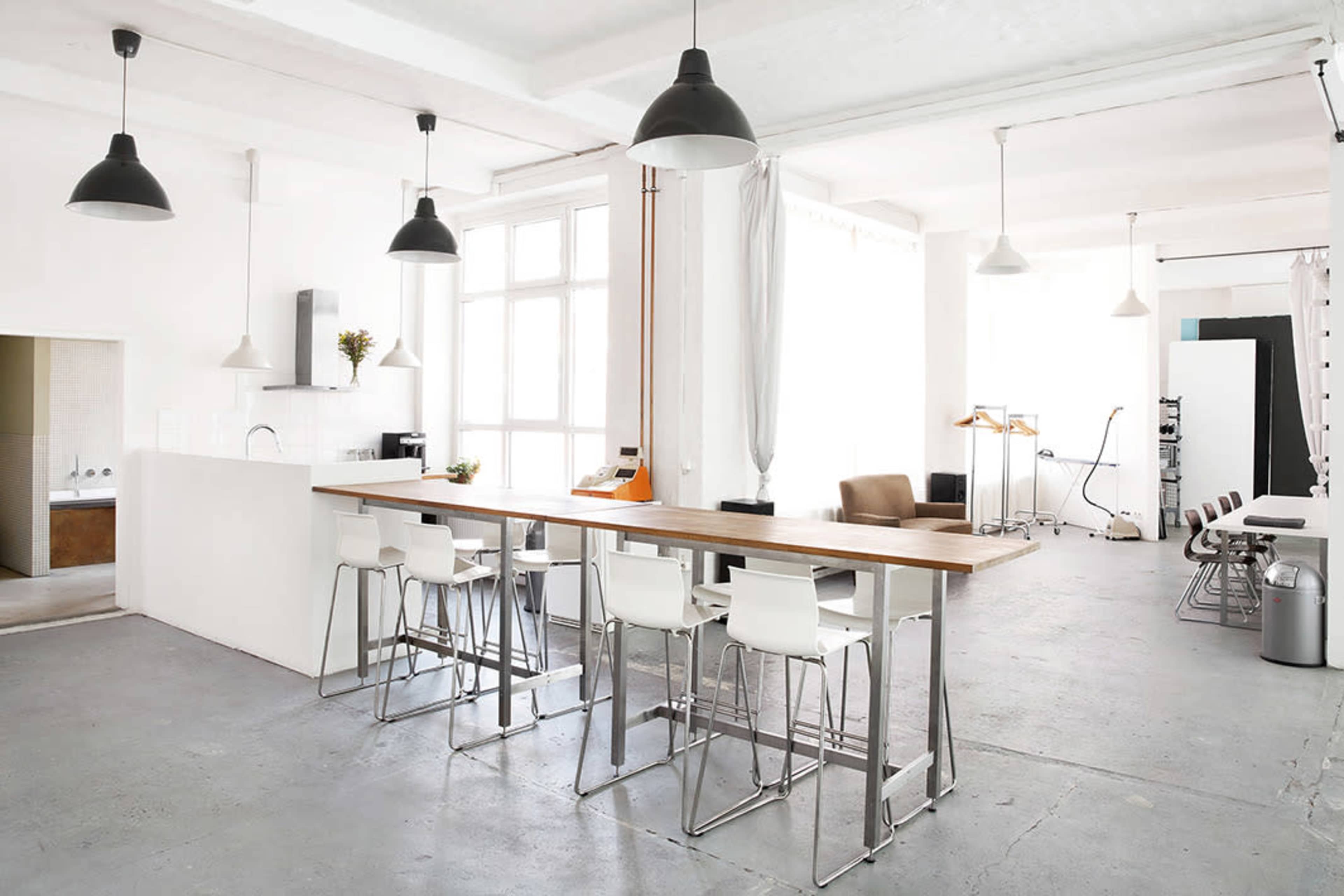 A spacious, modern kitchen area with a long wooden bar and white stools, surrounded by bright walls and industrial lighting.