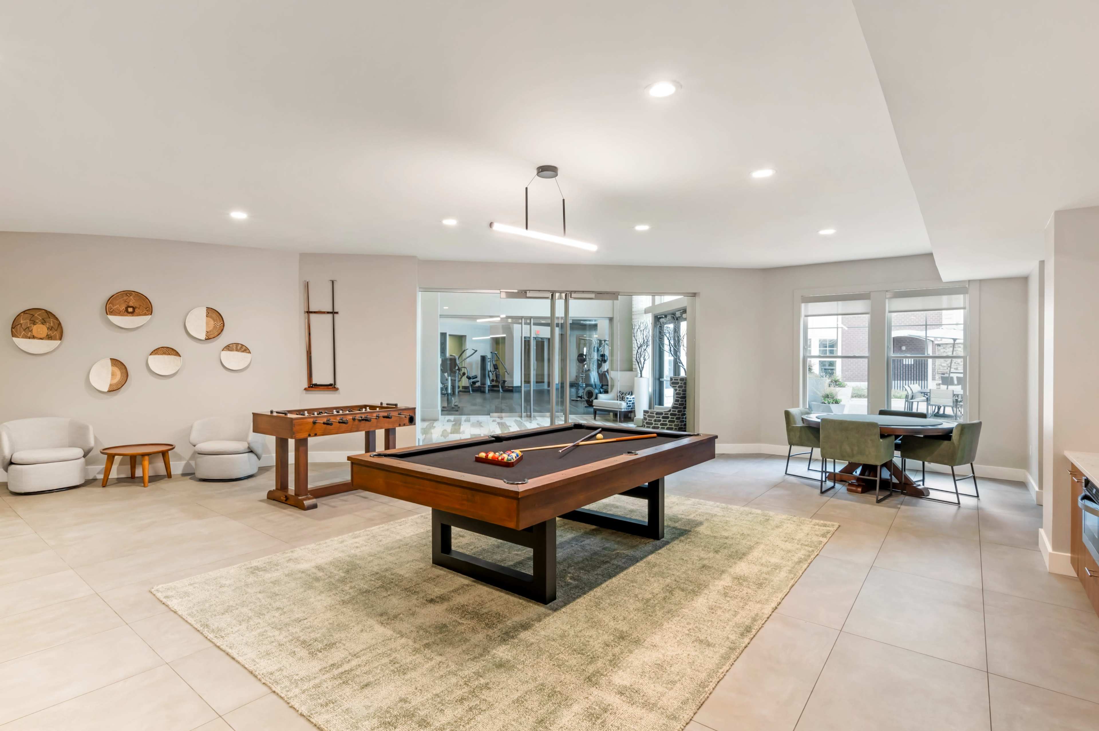 The image shows a spacious recreational room featuring a pool table, a foosball table, seating areas, and large windows that lead to an outdoor space.