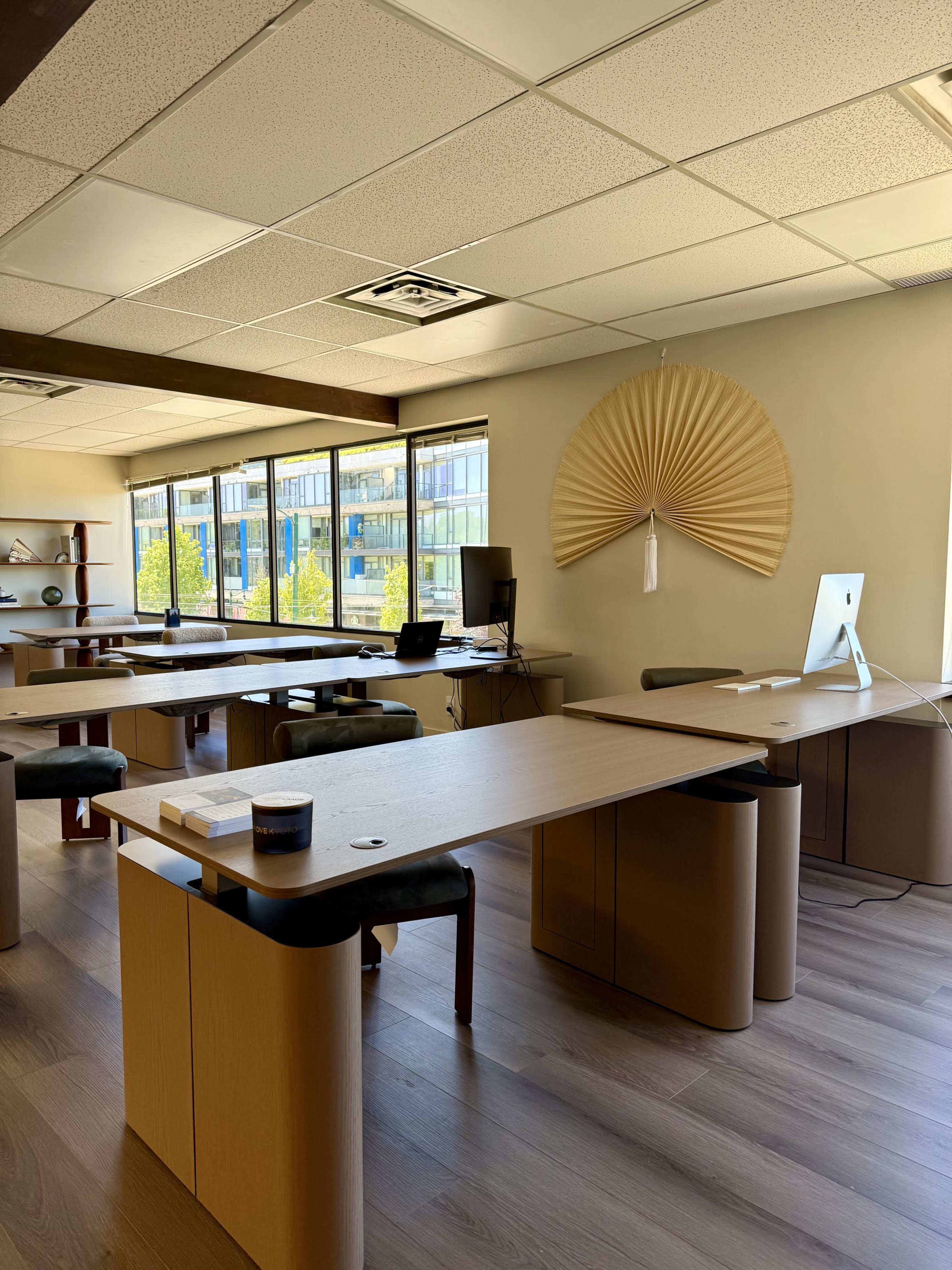 The image shows a modern office space with multiple desks, large windows, and a decorative fan on the wall.