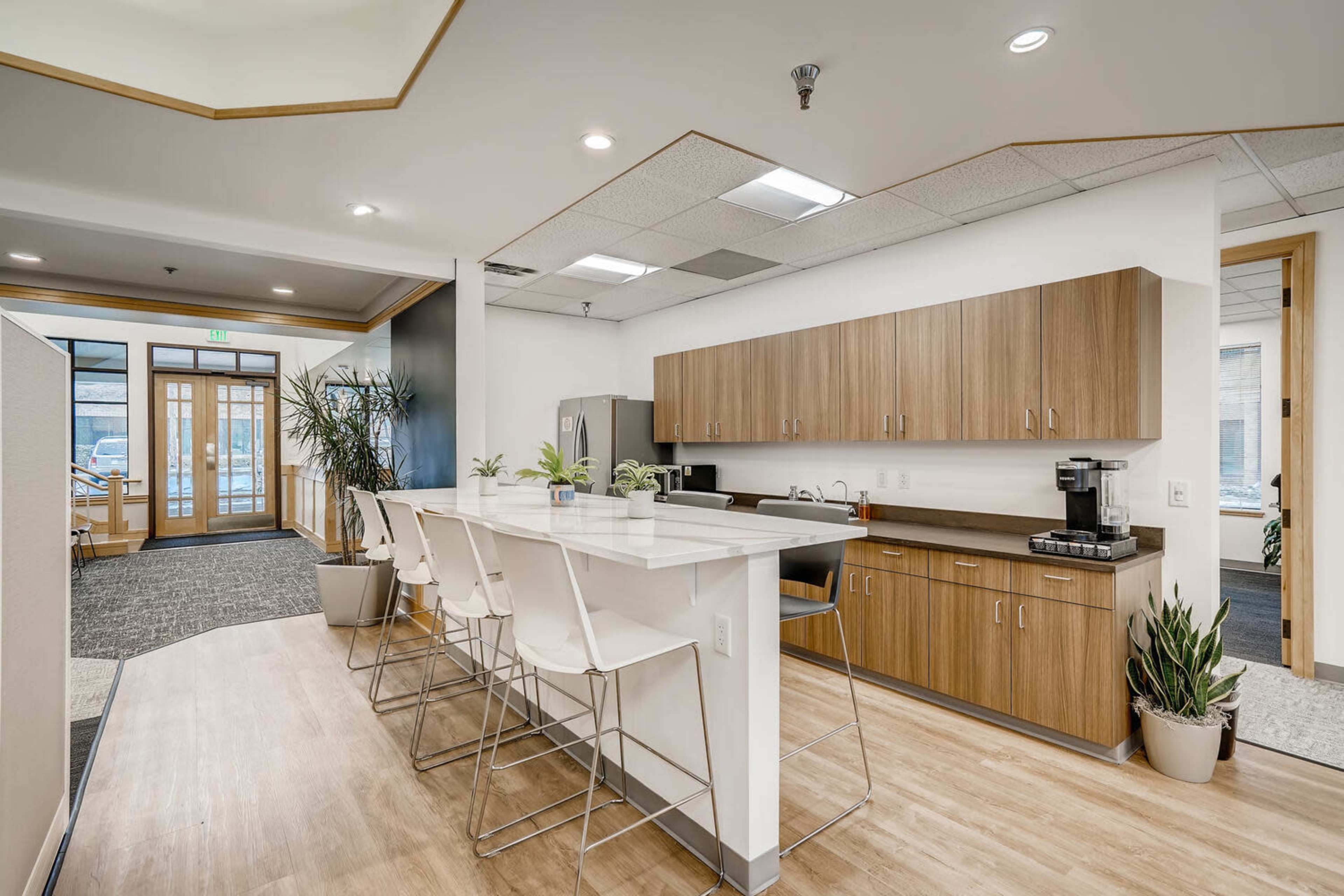 A modern kitchen area with wooden cabinets, a white countertop, and high stools, surrounded by potted plants.