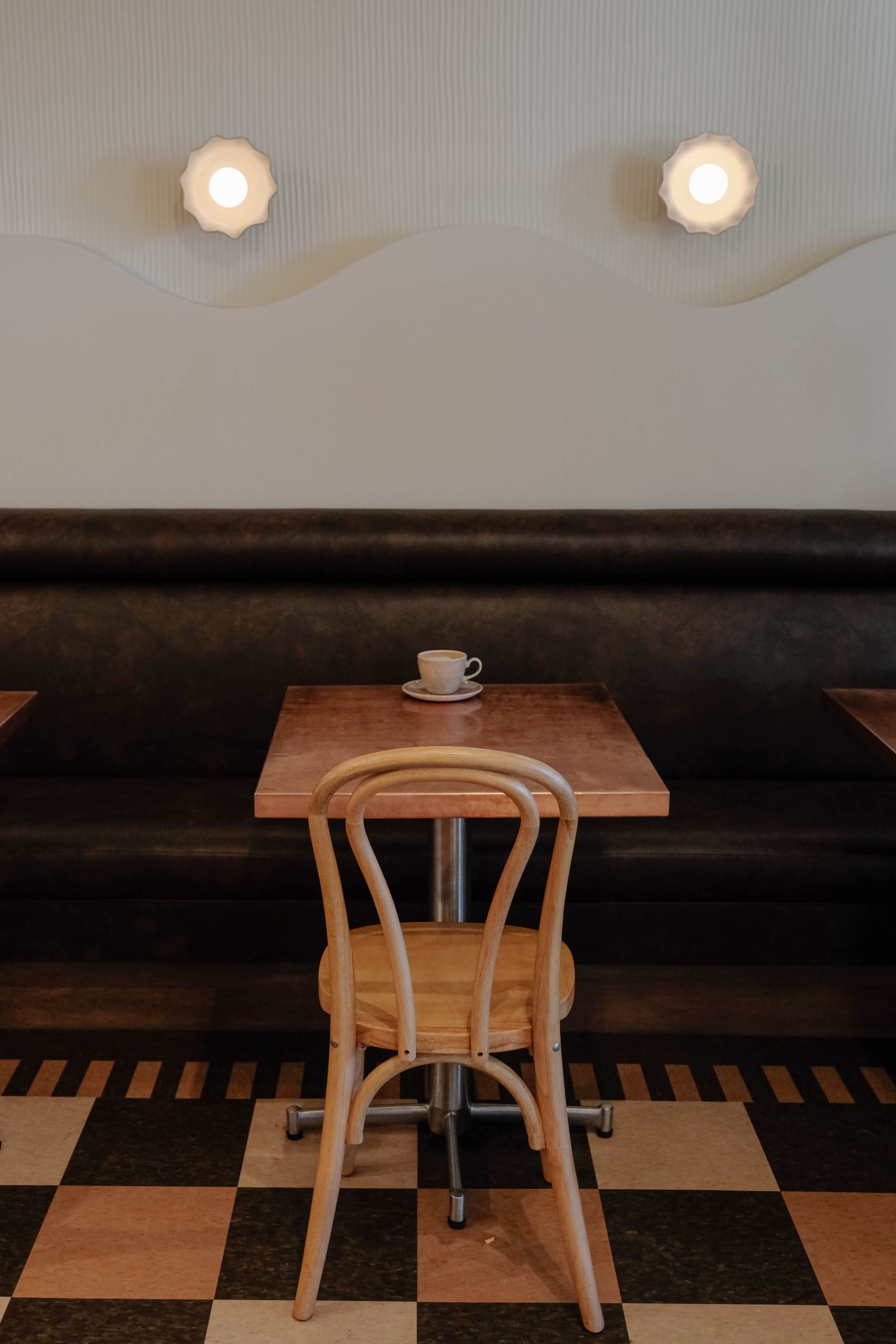 A single wooden chair is positioned next to a small table with a coffee cup on it in a cafe featuring simple wall lights and a patterned floor.