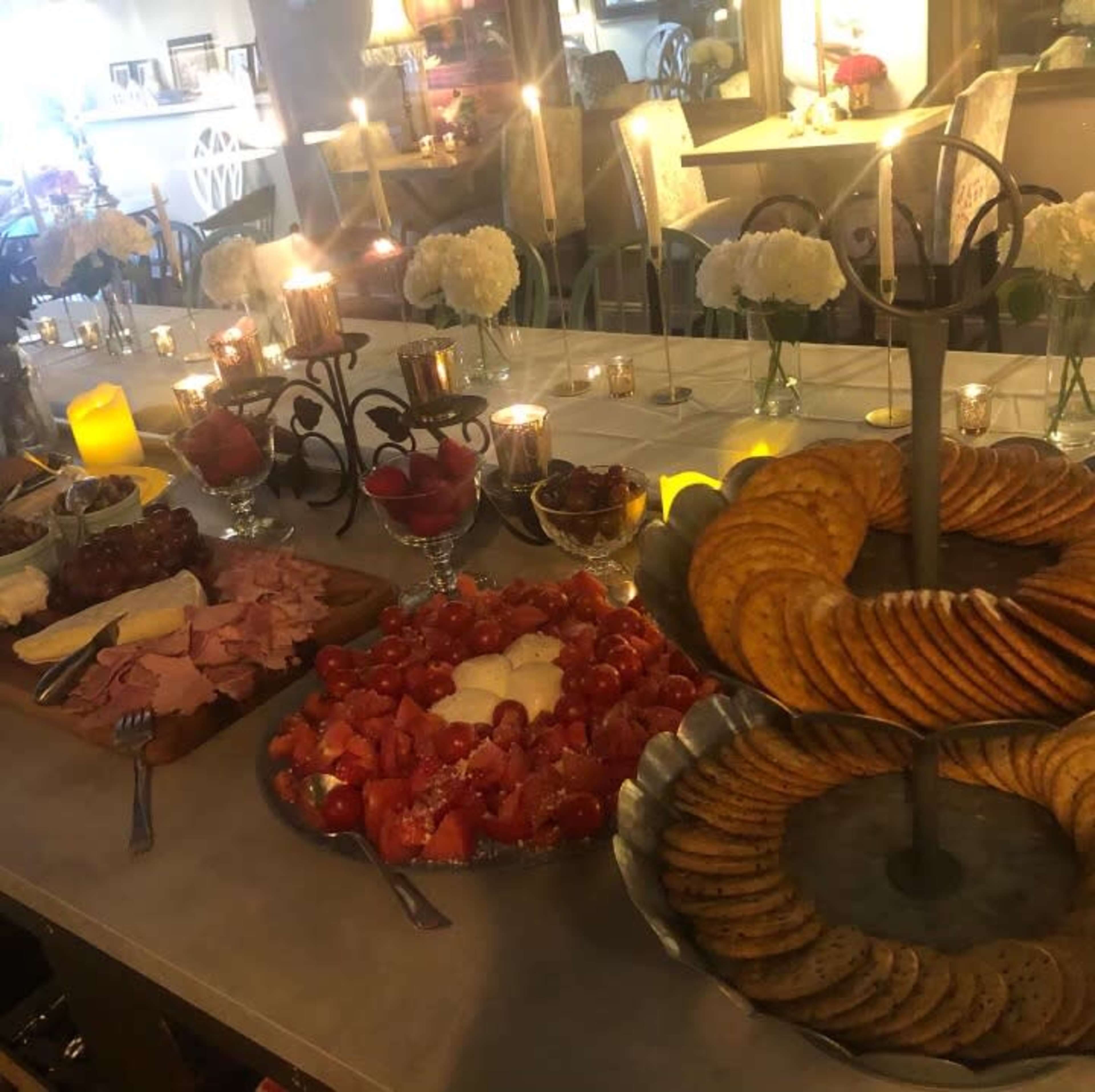 The table is set with various food items, including sliced meats, cheese, fresh tomatoes, cookies, and fruit, along with lit candles and floral arrangements.