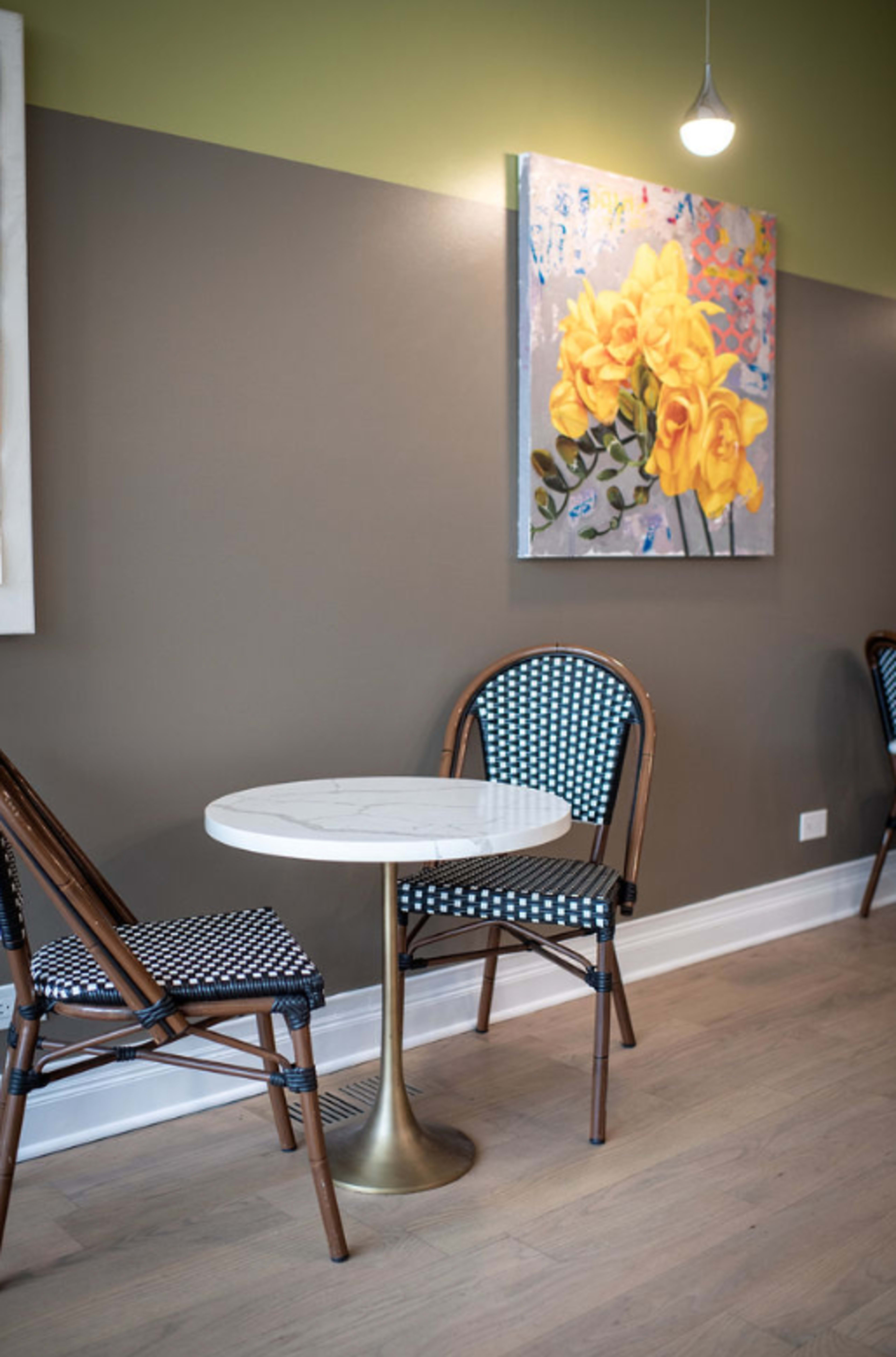 A small round marble table is set between two patterned chairs against a wall adorned with a floral painting.