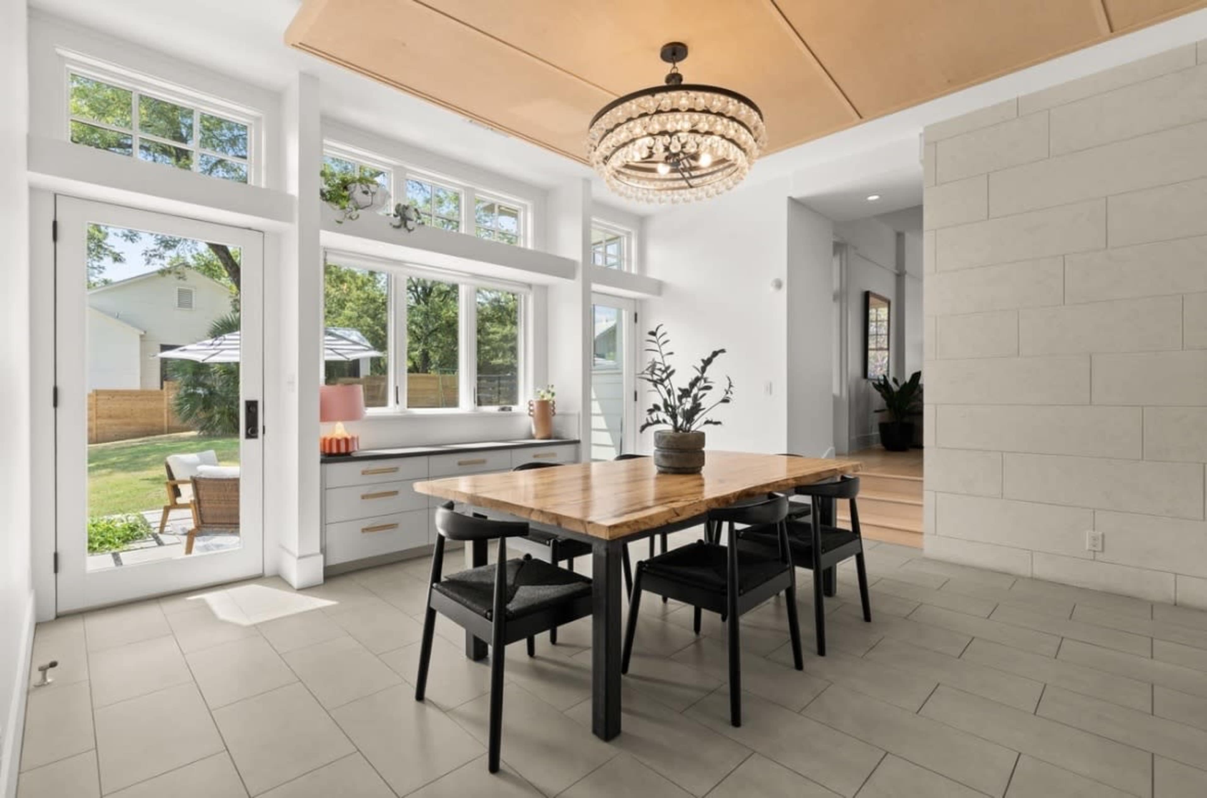 A modern dining area features a wooden table surrounded by black chairs, with large windows overlooking a backyard and a chandelier overhead.