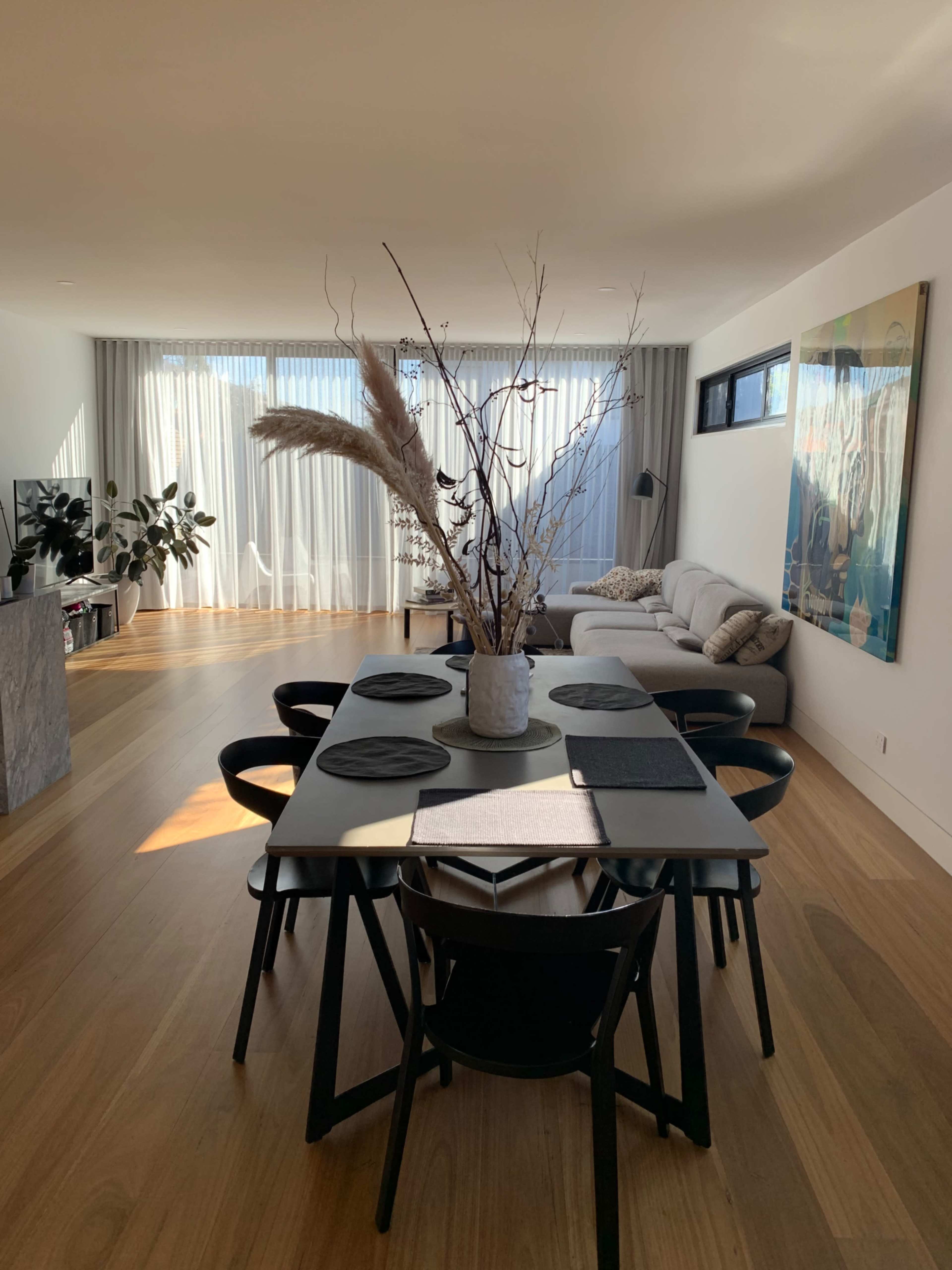 A modern dining room features a table with black chairs, a tall decorative vase with branches, and large windows allowing natural light to fill the space.