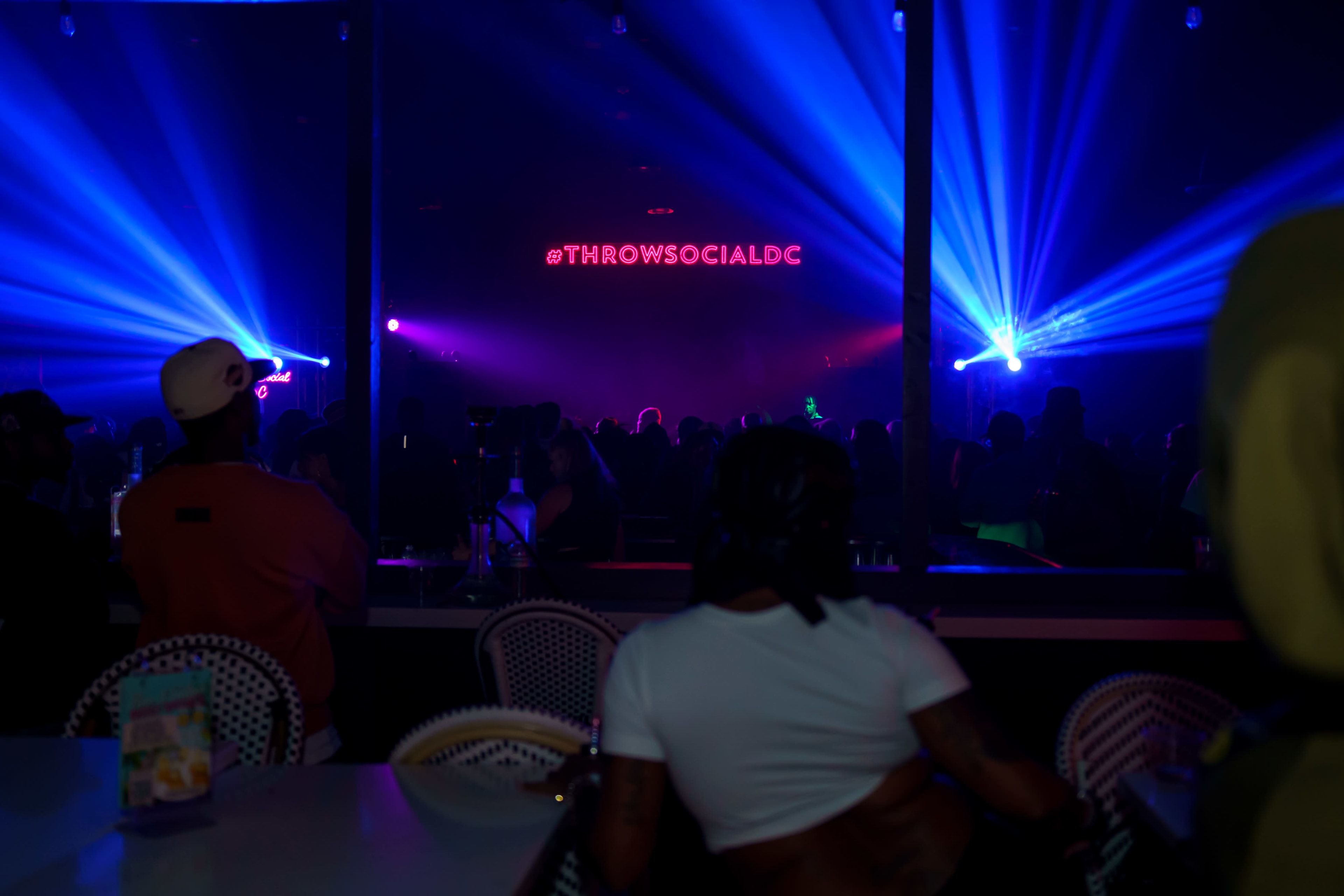 A crowded venue with a stage illuminated by colorful lights, featuring the text "@THROWSOCIALDC" in the background.