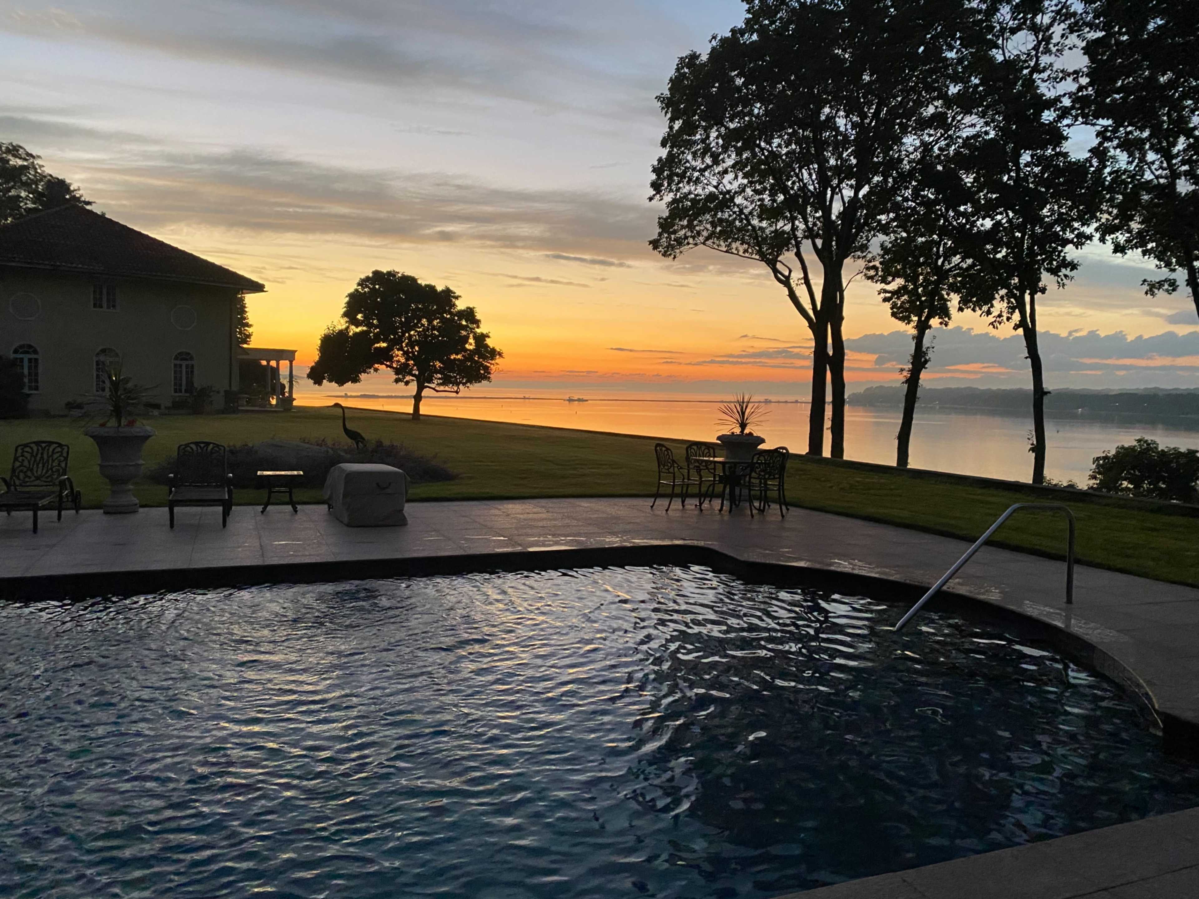 A swimming pool reflects the colors of a sunset over a calm lake, with a house and trees in the background.