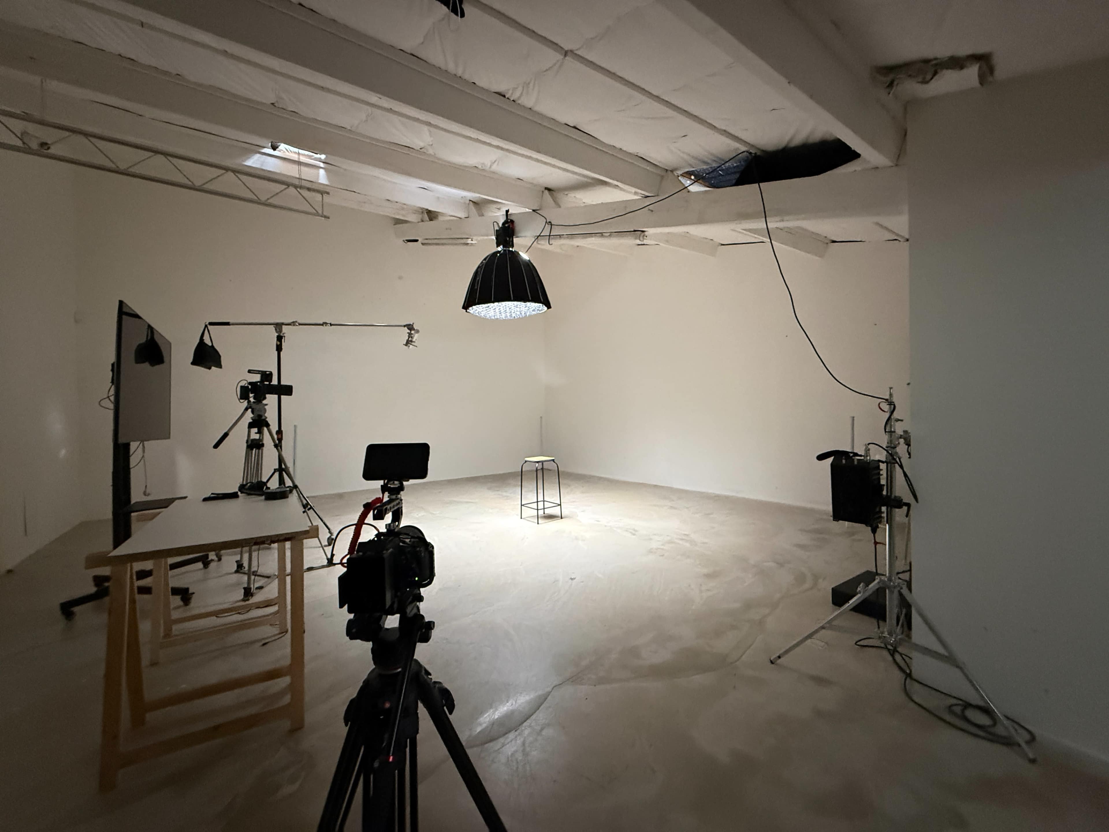 The image shows a minimally furnished photography studio with a single stool in the center, surrounded by various lighting equipment and cameras.