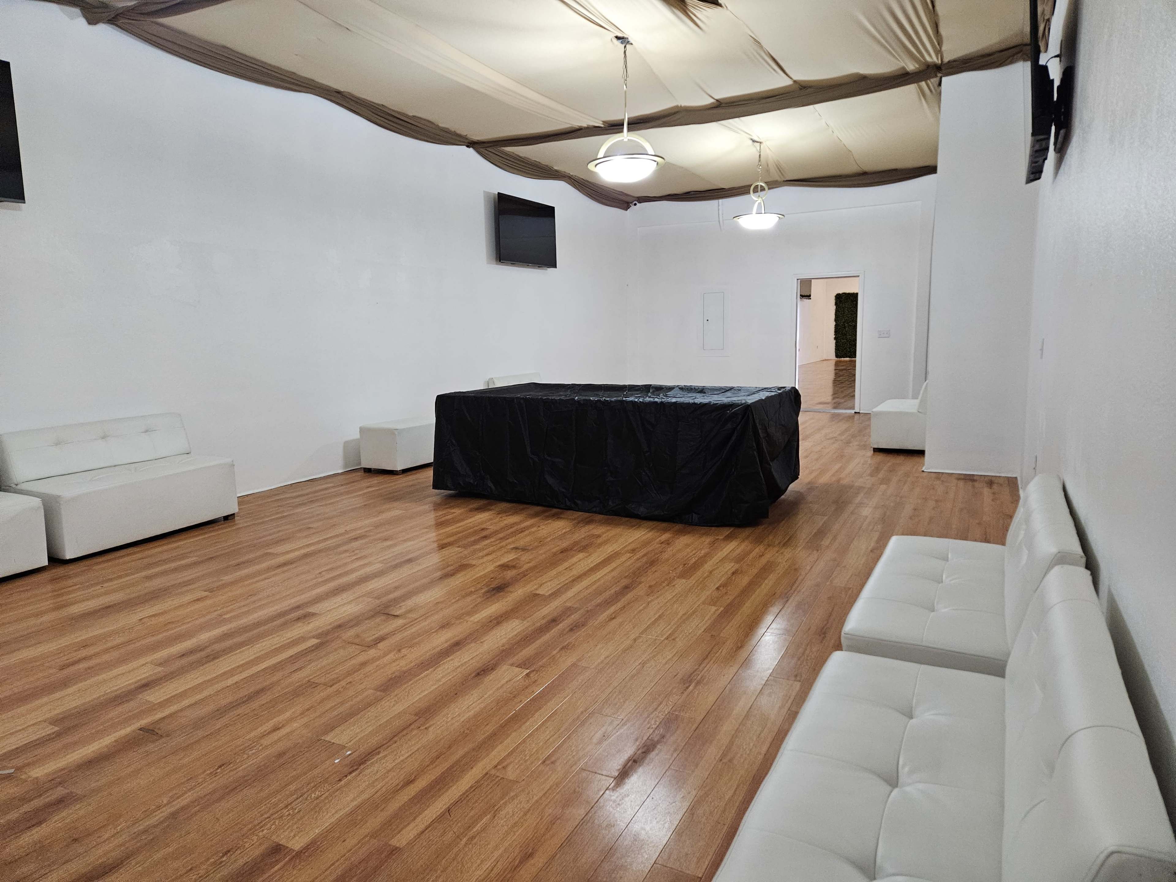 The room features white walls and a wooden floor, with a large black-covered table in the center, surrounded by white couches and equipped with two wall-mounted screens.