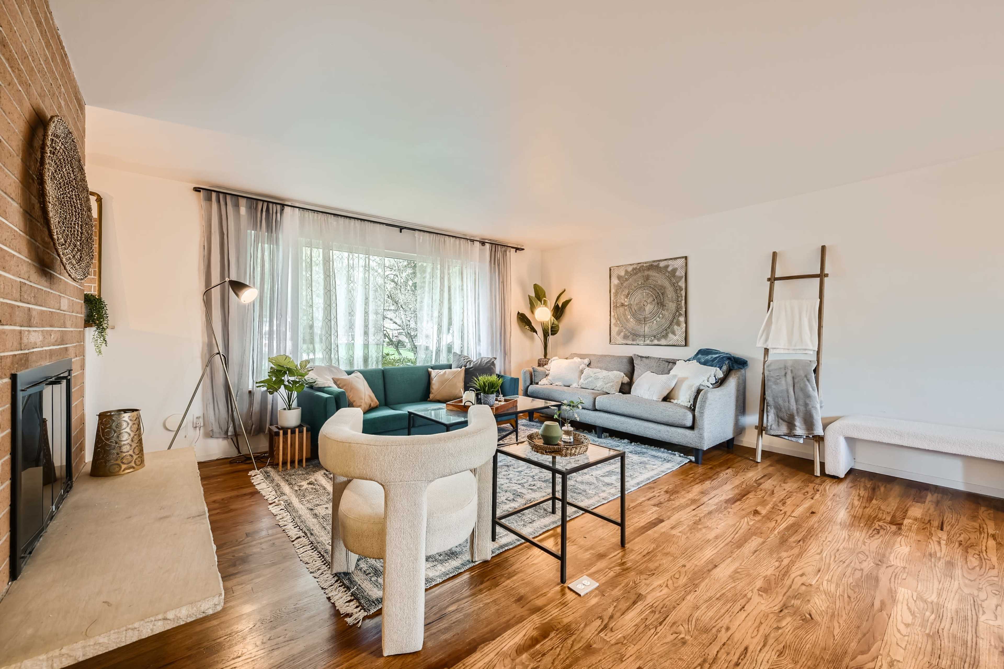 The image shows a modern living room with a sectional sofa, a round coffee table, a light-colored rug, a fireplace, and large windows allowing natural light to enter.