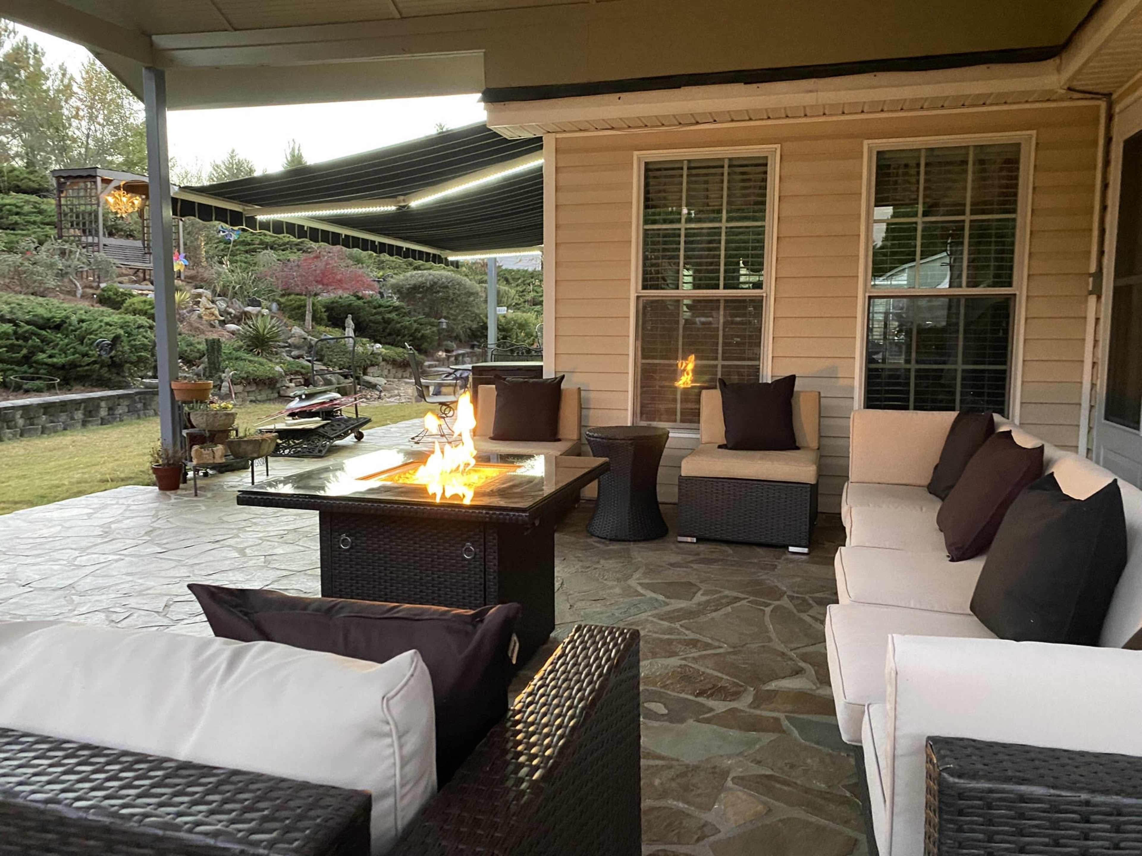 A patio area featuring a fire pit table surrounded by outdoor seating and a landscaped yard in the background.