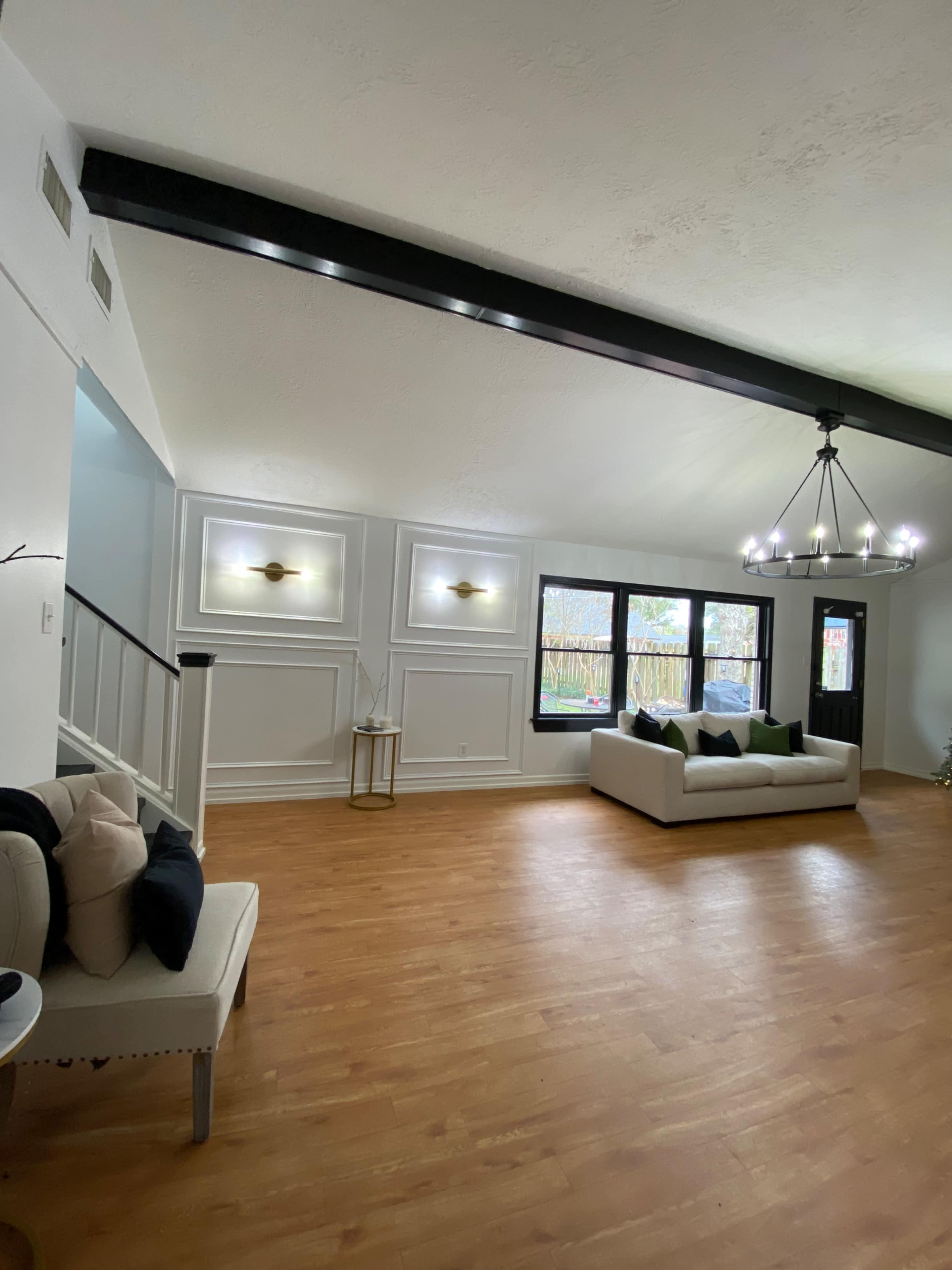 A spacious living room features a high ceiling with dark beams, large windows, and a light-colored sofa surrounded by decorative pillows.