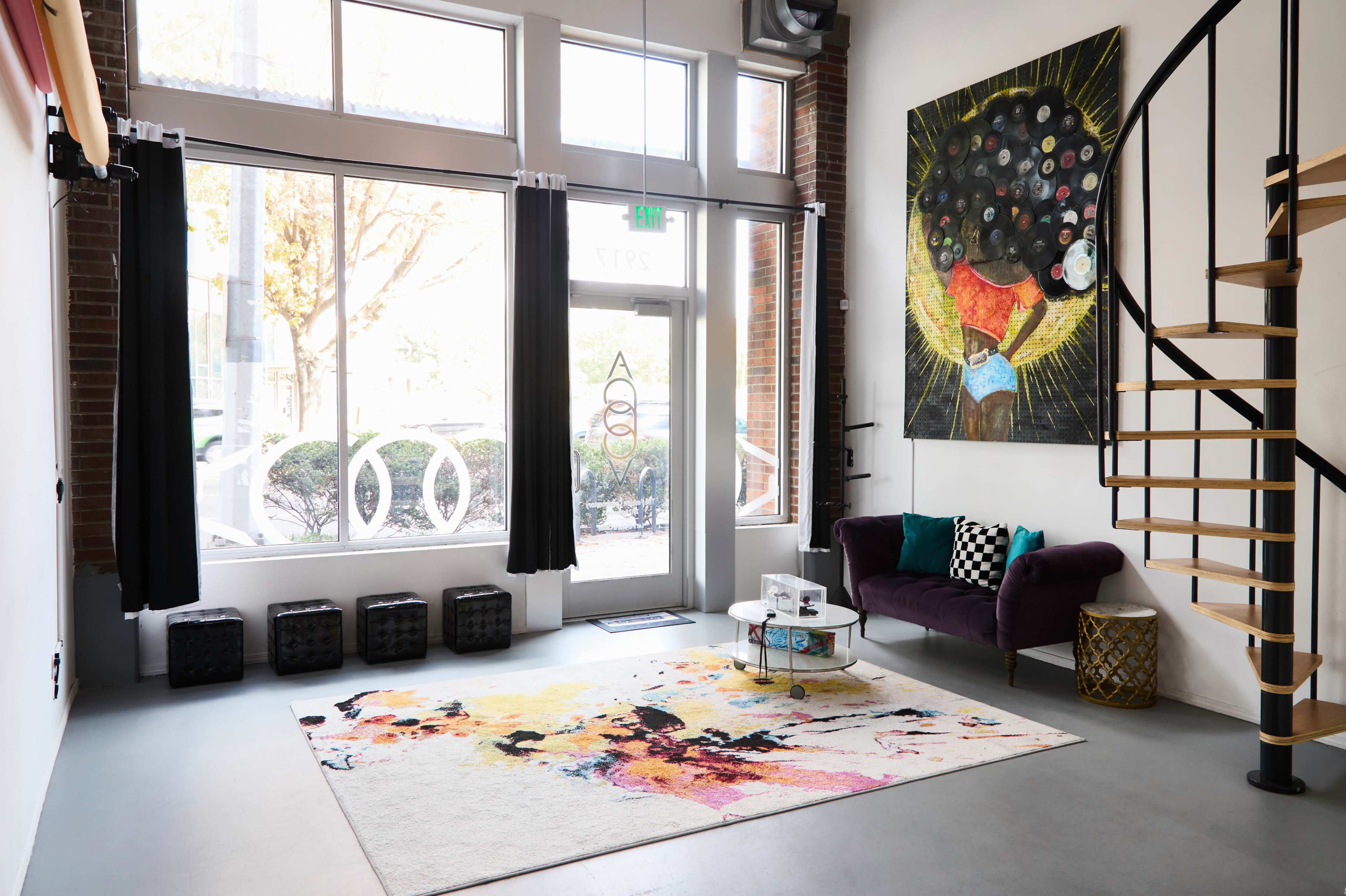 The image shows a modern interior with large windows, a spiral staircase, a colorful rug, a purple sofa, and vibrant wall art.