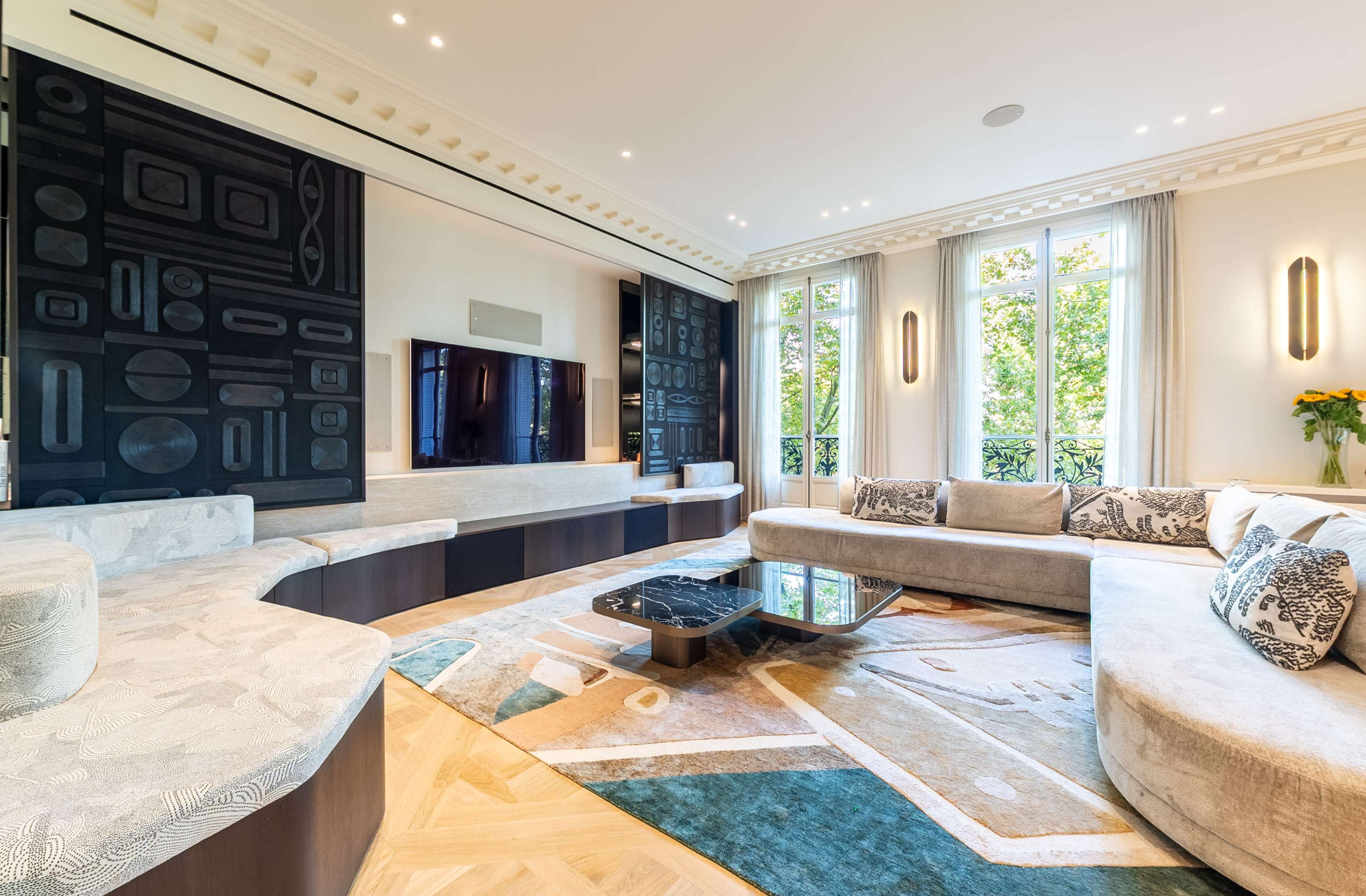 The image shows a modern living room featuring a curved sofa, a large television mounted on a dark wall, and ample natural light from large windows.