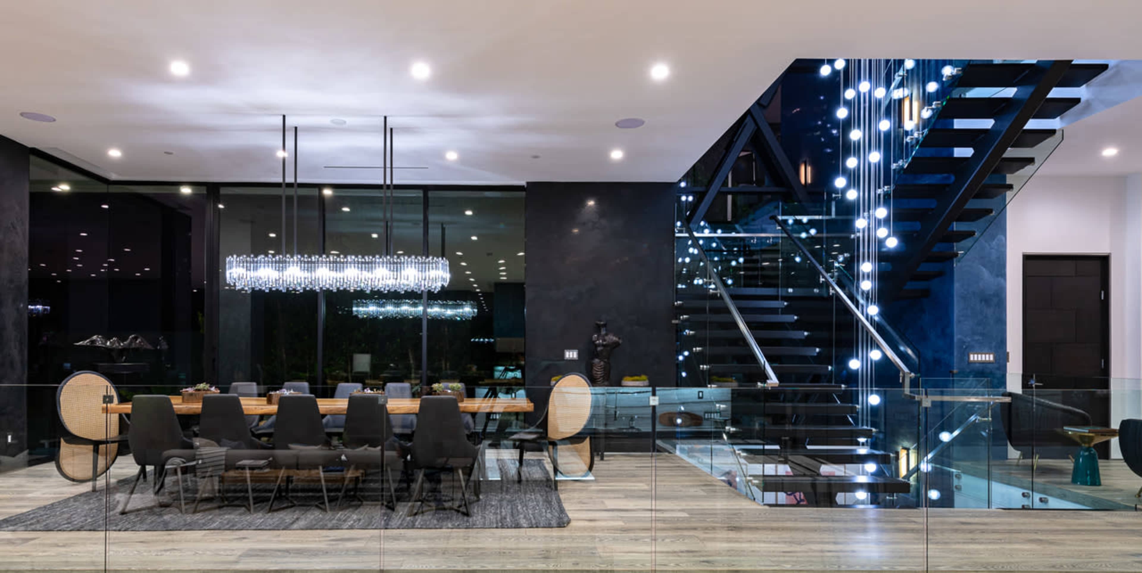 A modern dining area with a long table surrounded by chairs, adjacent to a staircase illuminated by decorative lights.