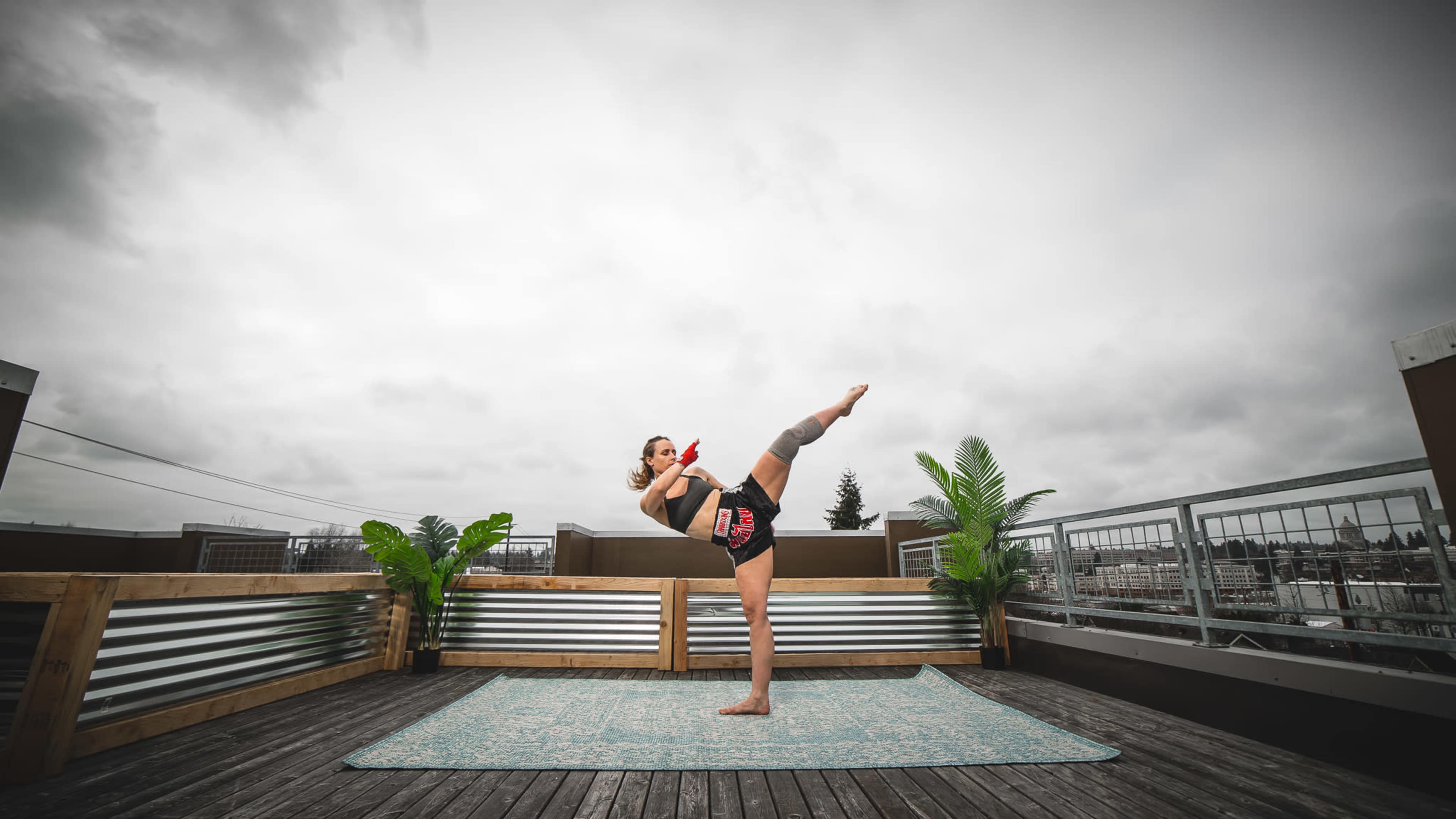 A person performs a high kick on a rooftop deck with potted plants and a gray sky in the background.