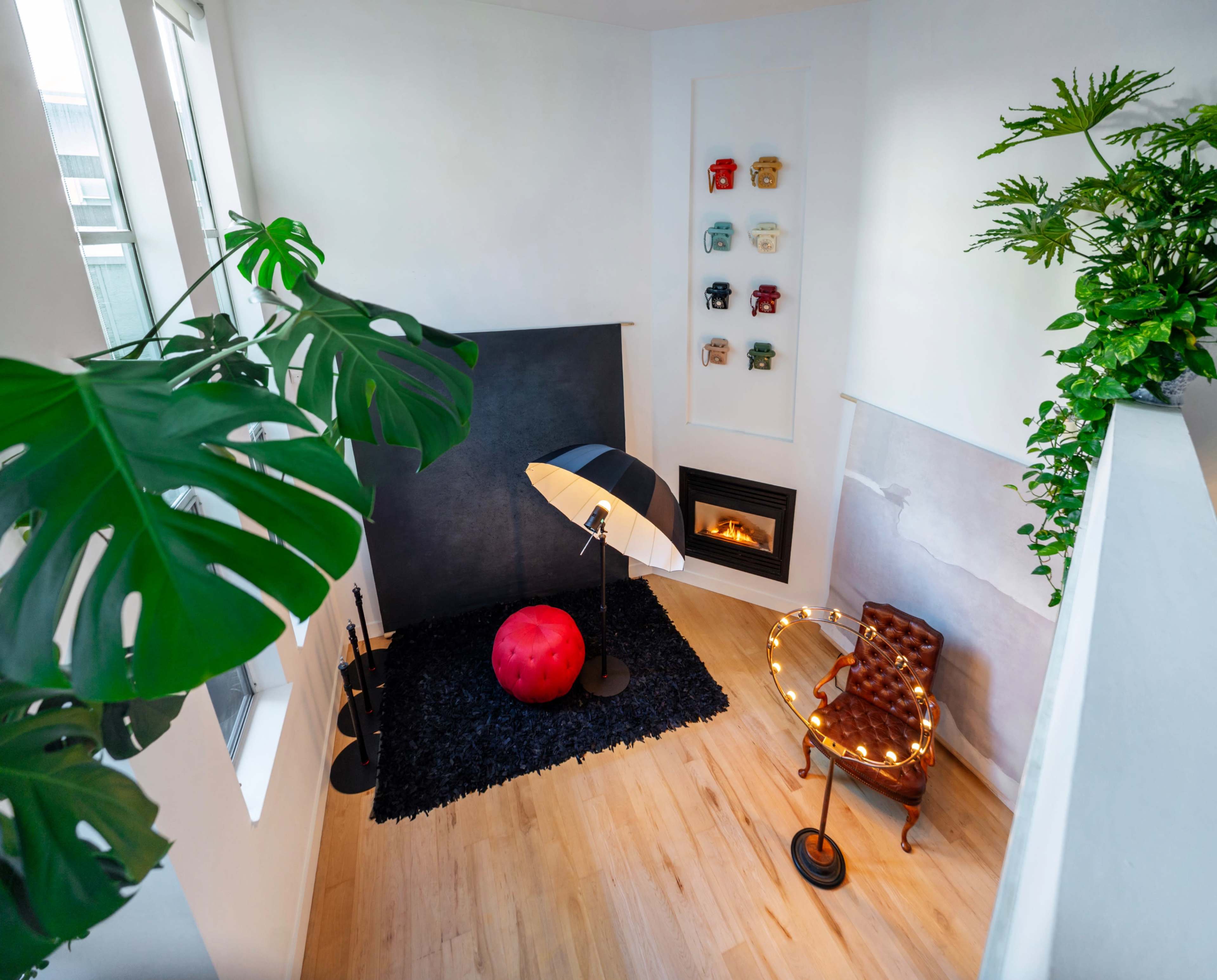 The image shows a modern, minimalist corner of a room featuring a black floor lamp, a red circular rug, a chair with string lights, and a small fireplace against a white wall.