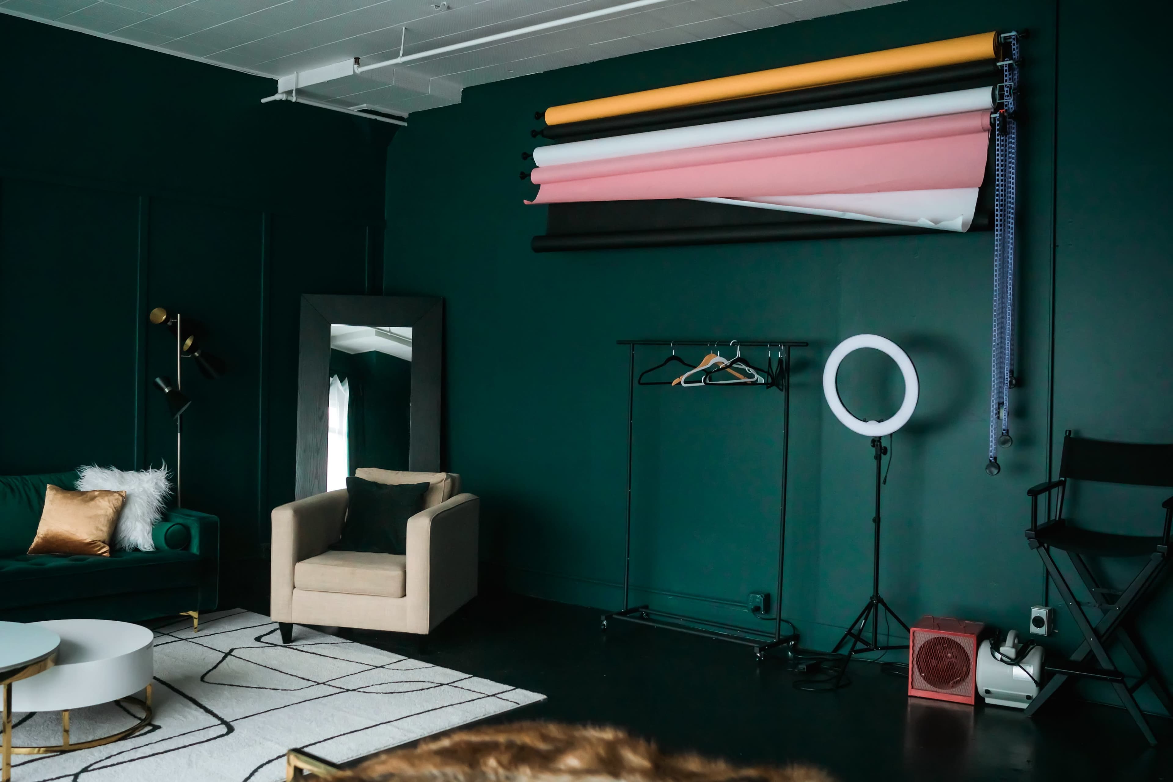 The image shows a modern studio space featuring a dark green wall, a large mirror, a clothing rack, and several rolls of backdrop paper arranged above.