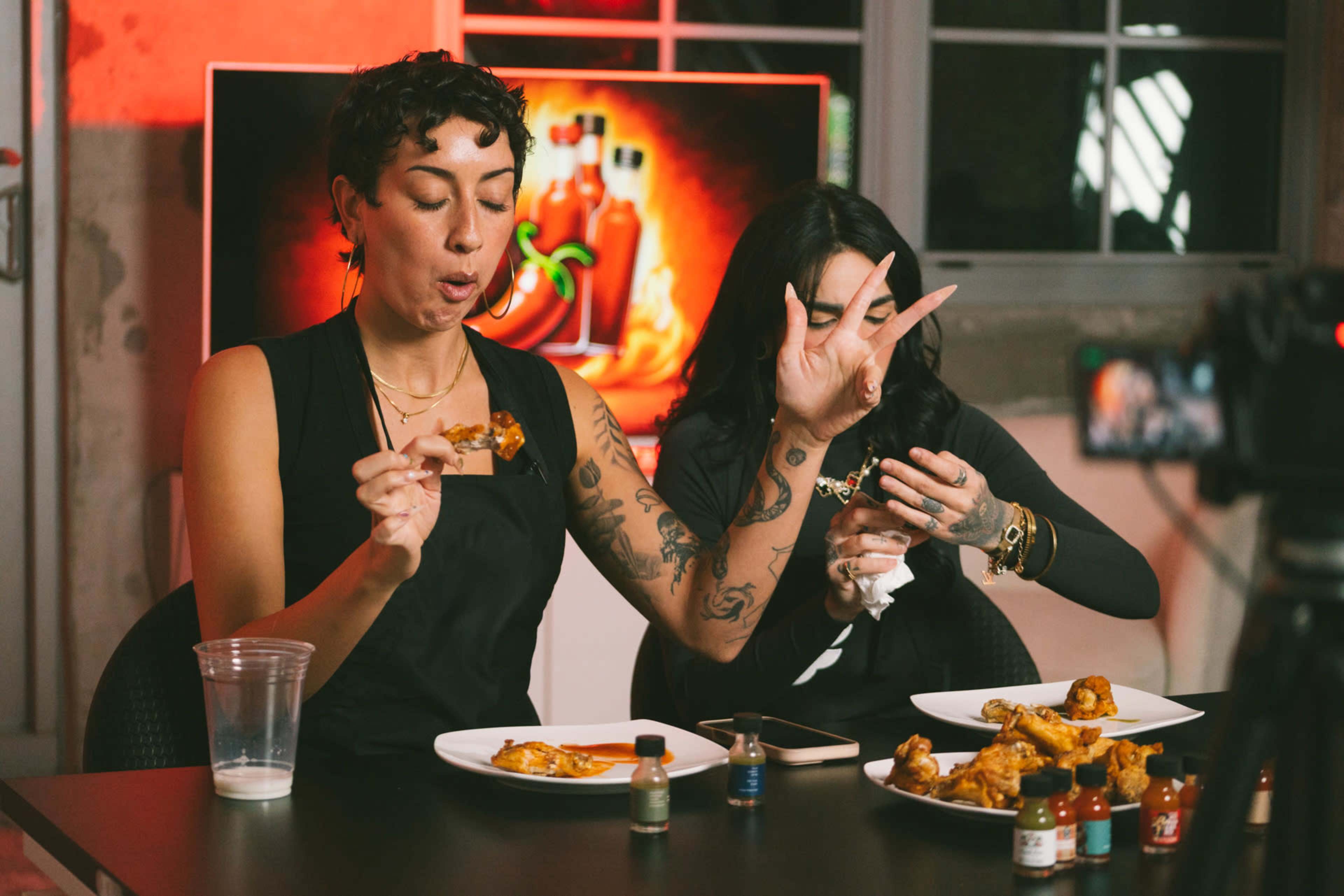 Two women are sitting at a table, eating chicken wings while various hot sauces are displayed in front of them.