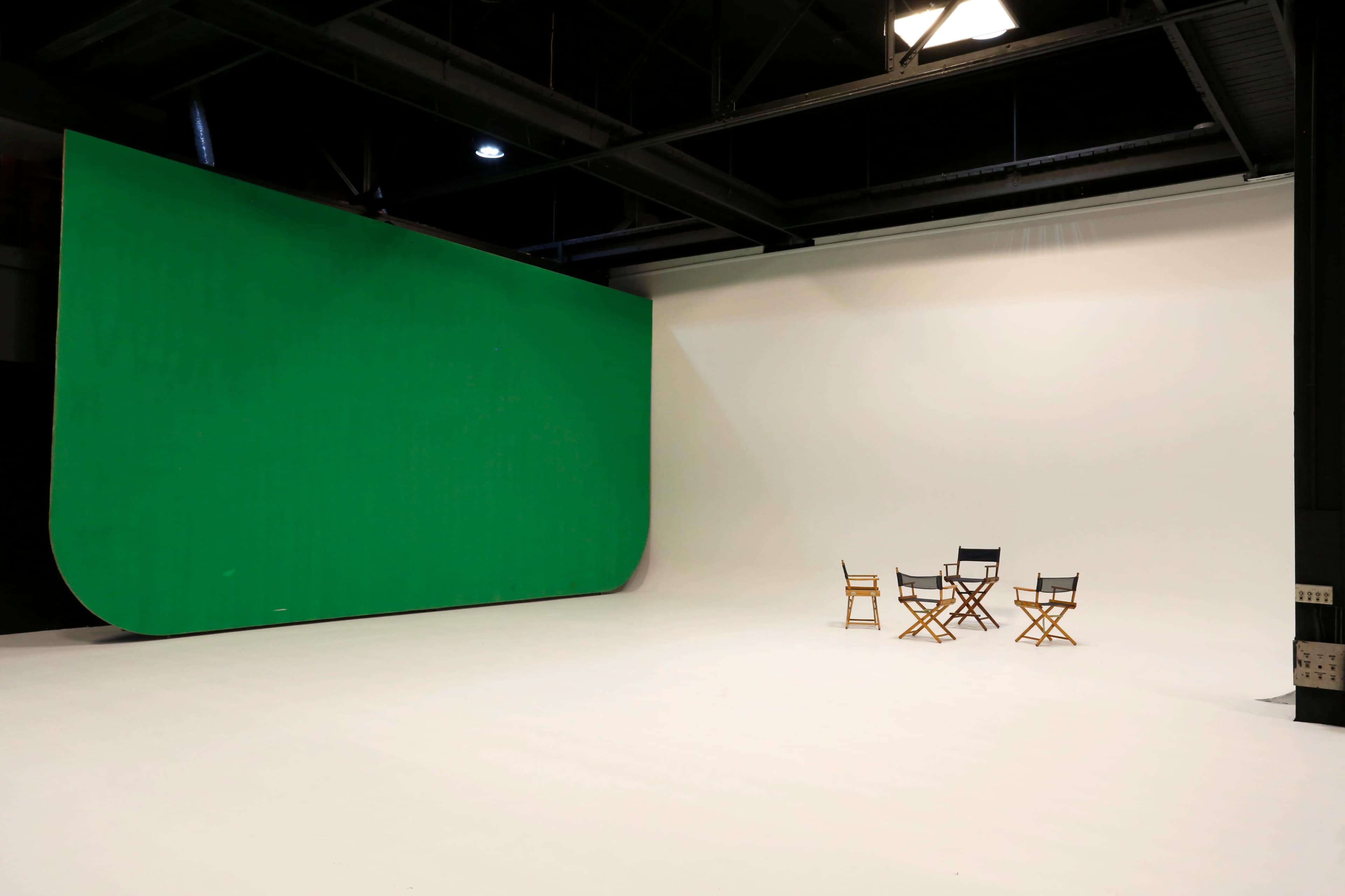 A studio space with a green screen on one wall and a set of three director's chairs arranged around a small table on a white floor.