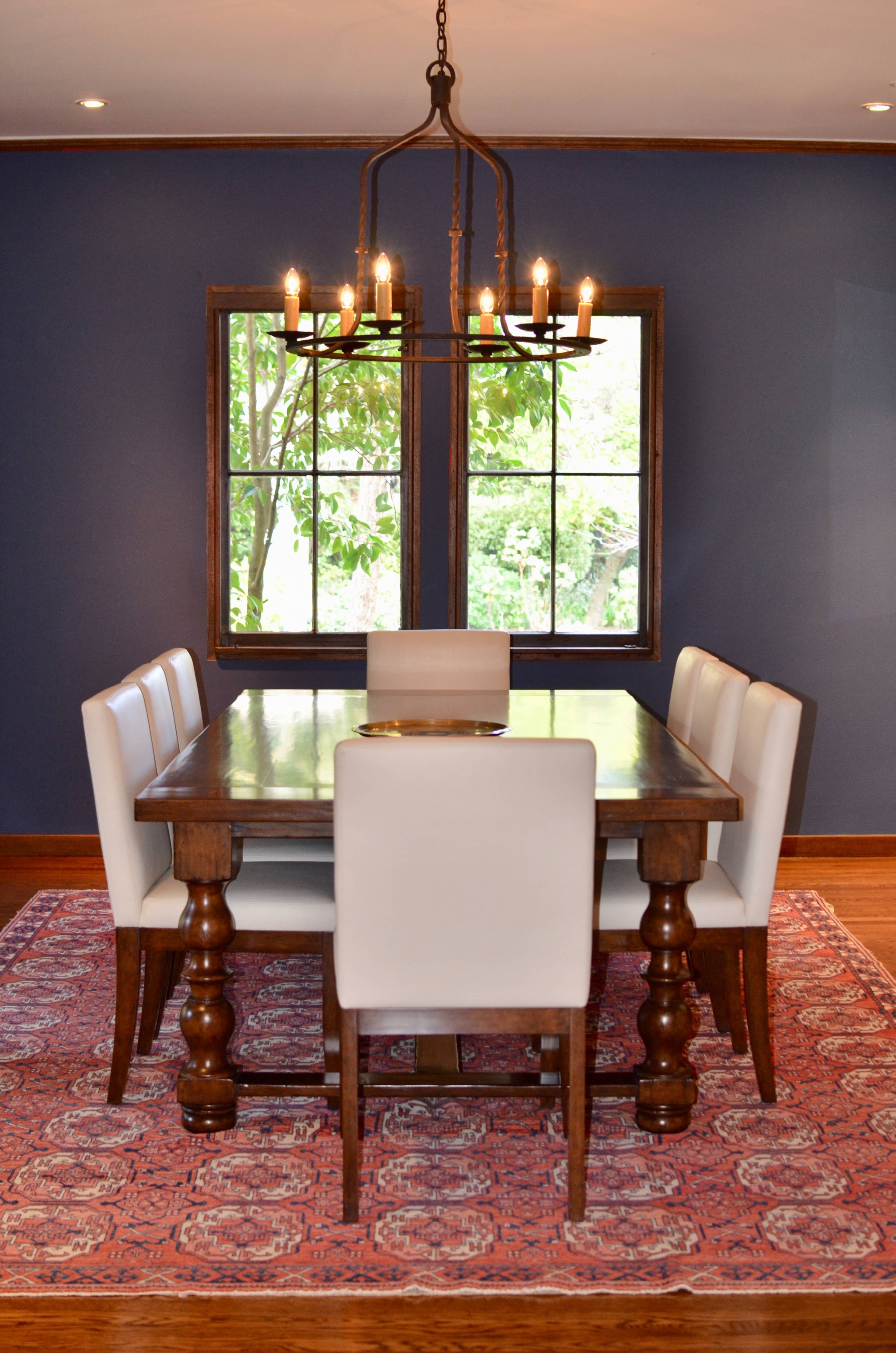 A wooden dining table is set with four white chairs on a patterned rug, under a chandelier, with two windows providing a view of greenery outside.