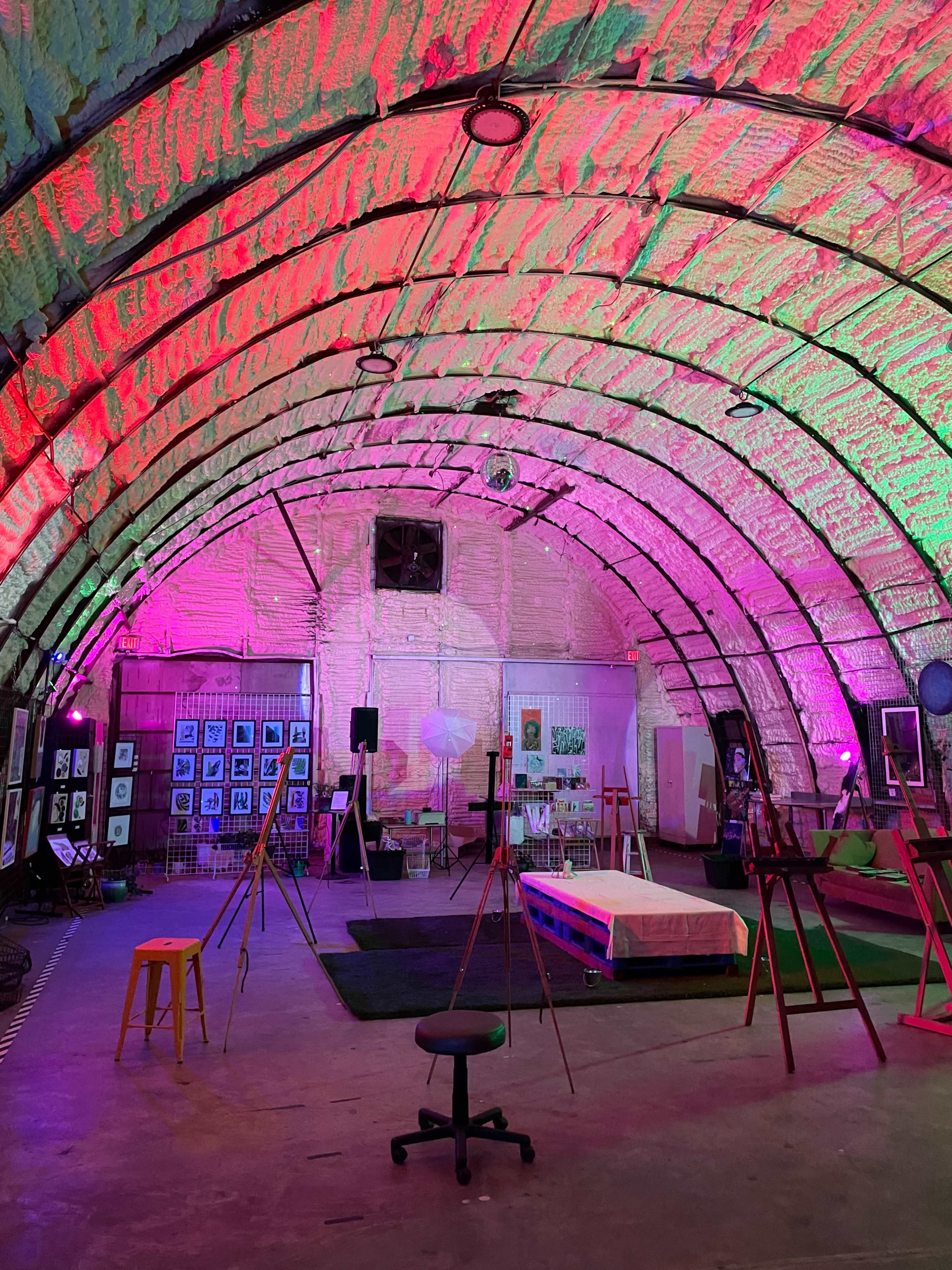 The interior of a large, dome-shaped space is illuminated with pink and green lighting, featuring art displays and setup for an event.