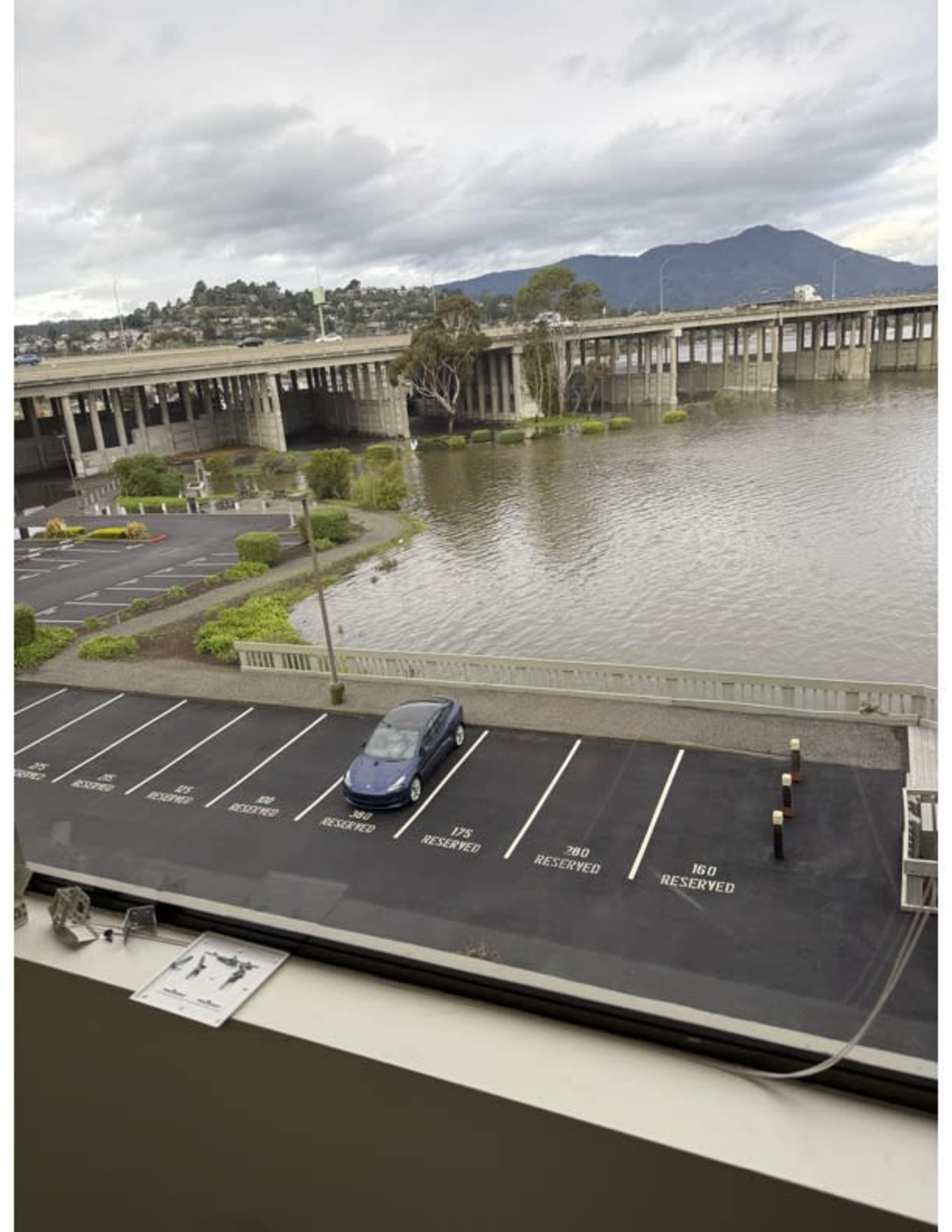 The image shows a flooded parking area adjacent to a river or body of water under a cloudy sky, with a single car parked in a space marked as reserved.