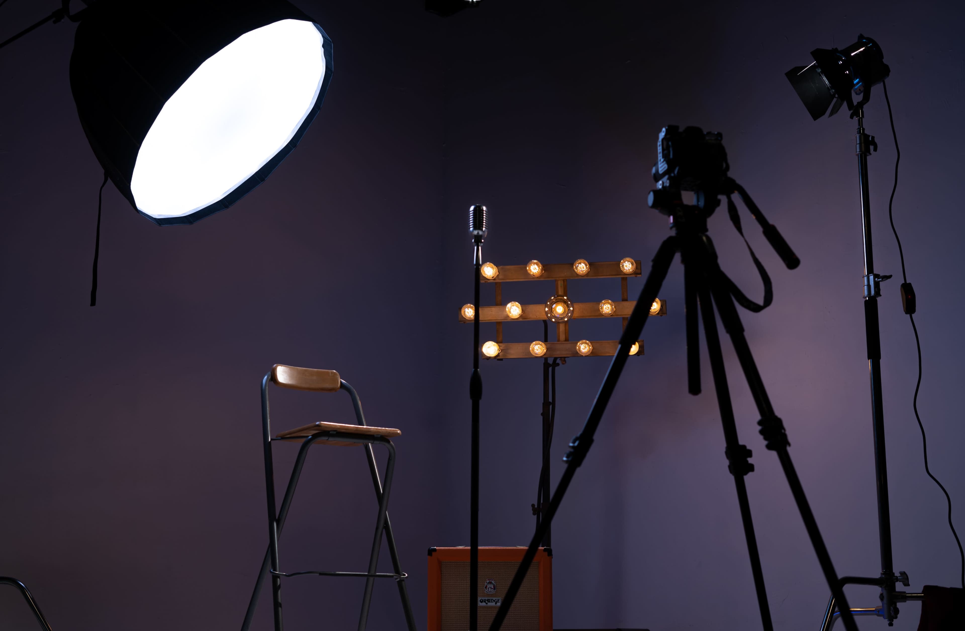 A photography studio setup features a chair, a microphone on a stand with lights, and a camera on a tripod against a dark purple wall.