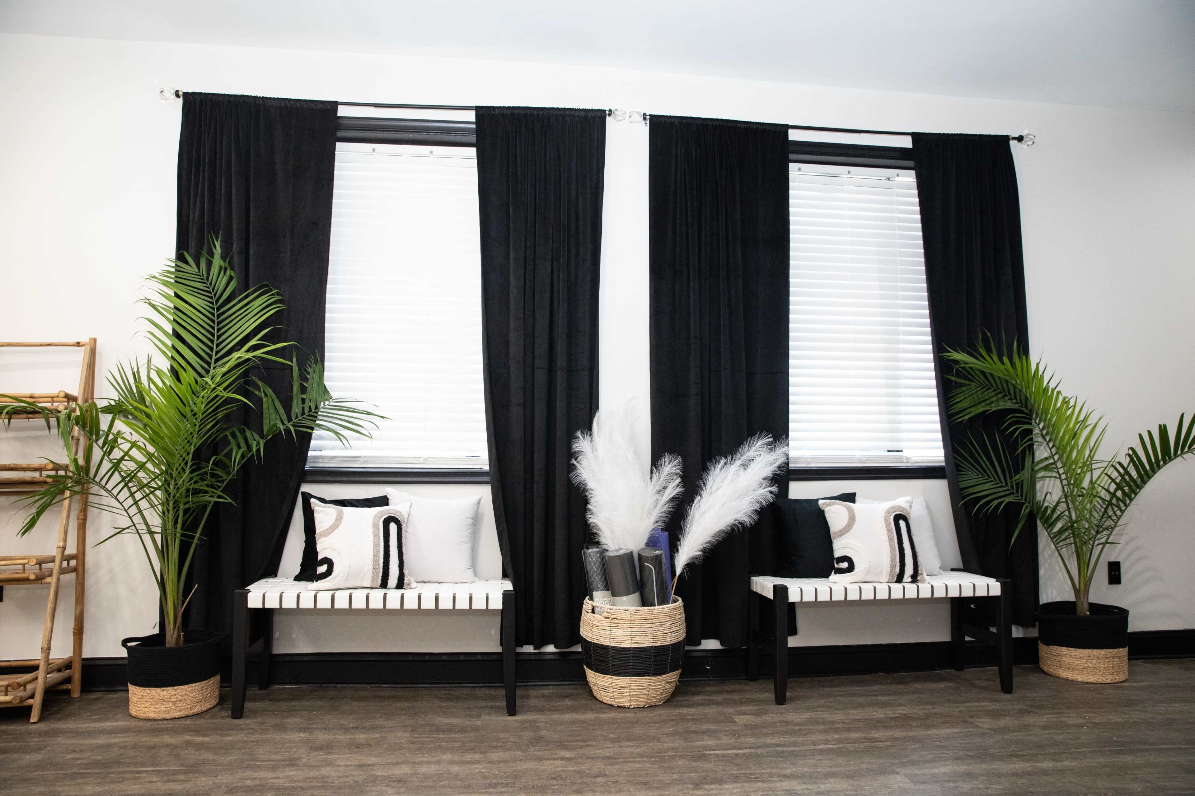 A room with two large windows dressed in black curtains, flanked by two benches and plants, with a shelf in the corner.