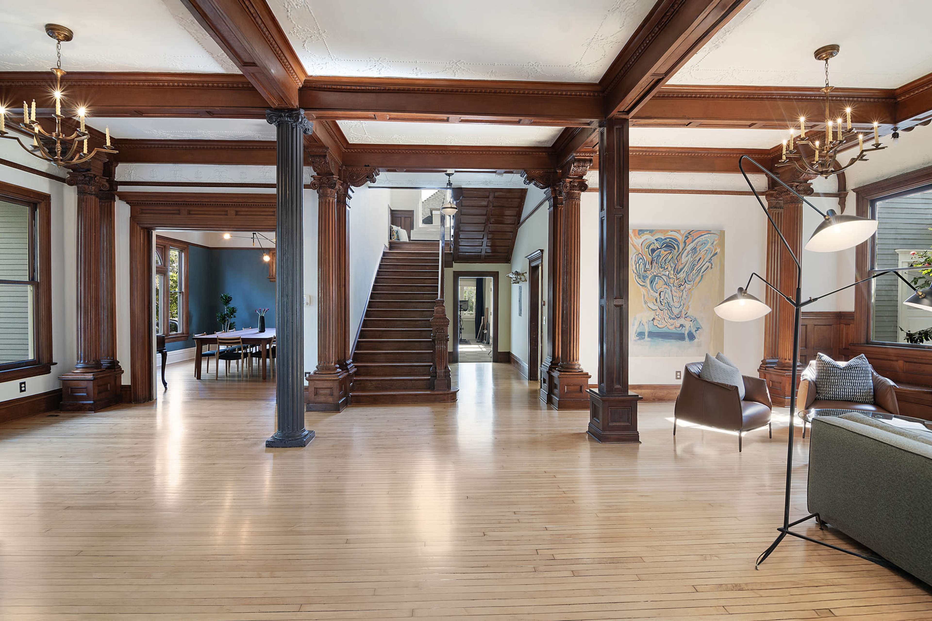The image shows a spacious interior with wooden pillars, a staircase in the background, and a combination of modern and traditional furniture.