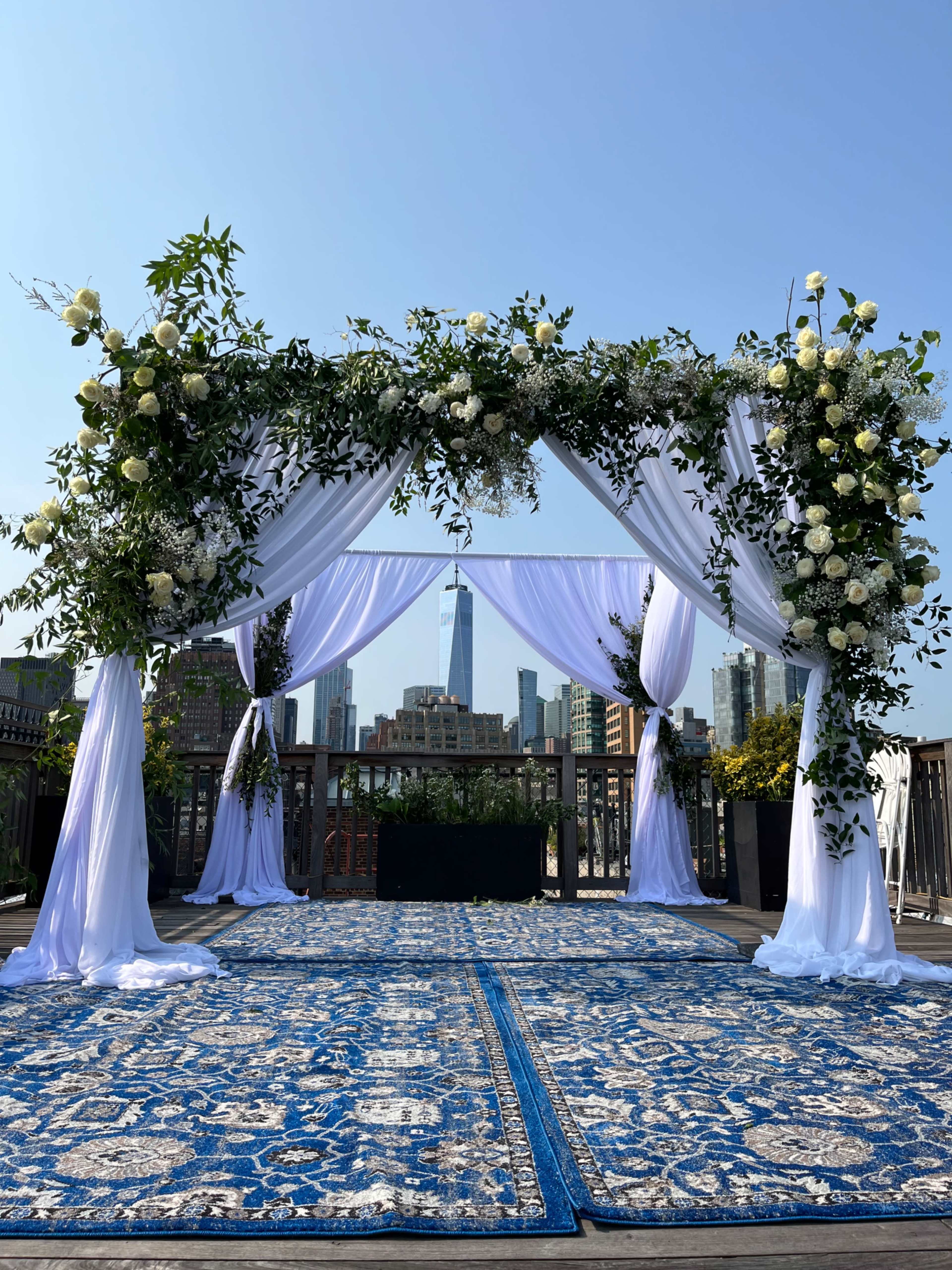 A wedding altar adorned with white drapes and floral arrangements, set against a skyline featuring a tall building in the background.
