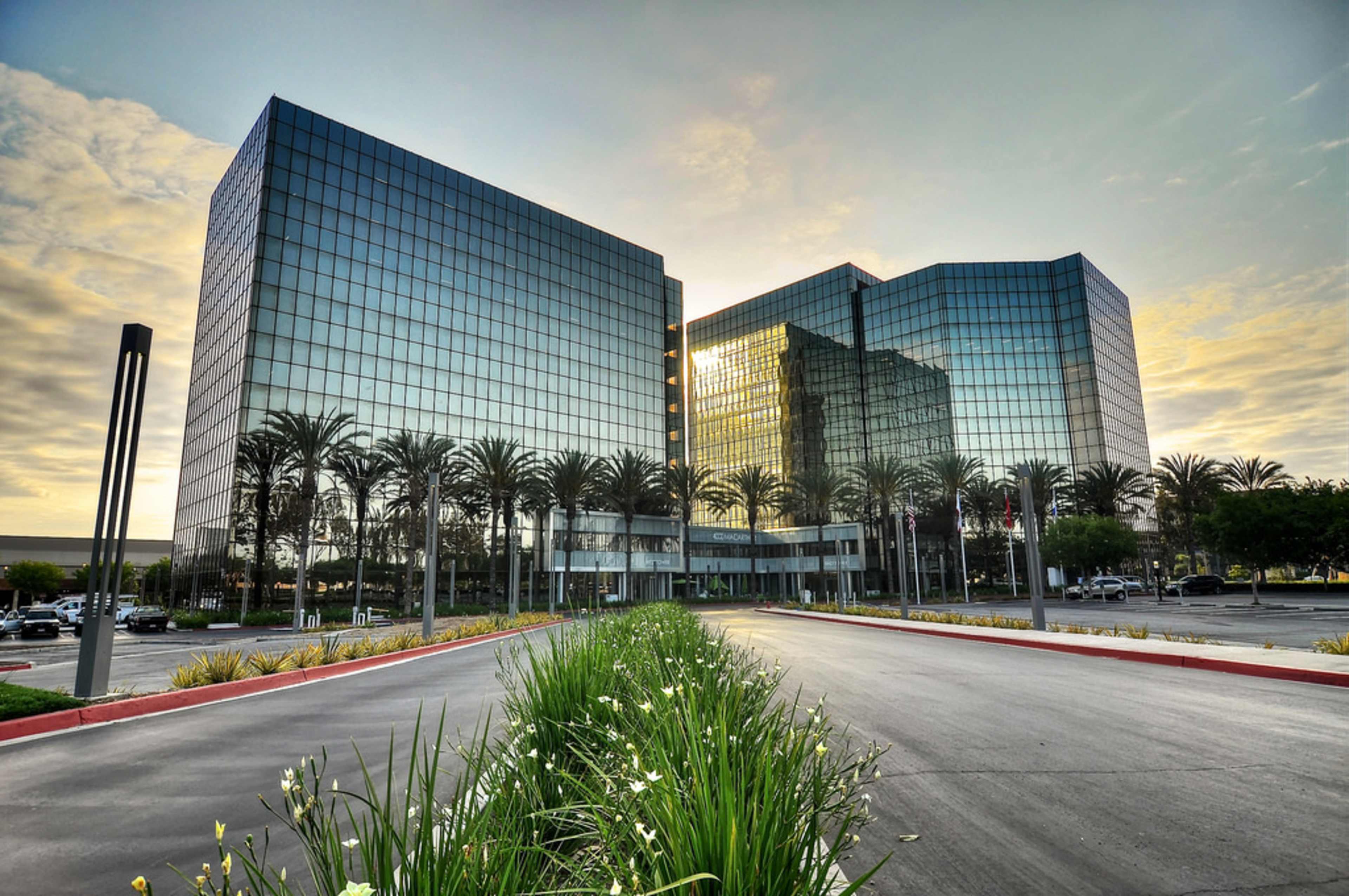 The image shows two glass-fronted office buildings flanked by palm trees and a landscaped path leading toward them.