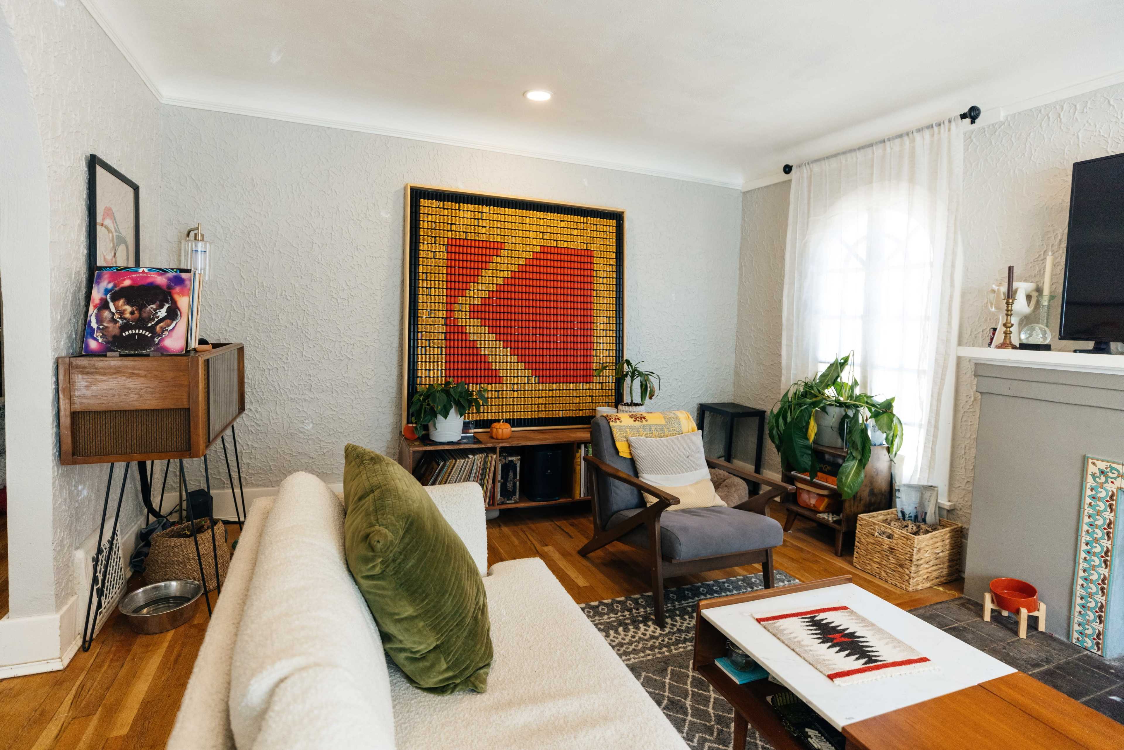 The living room features a couch, an armchair, a decorative wall with a large geometric artwork, and various plants.